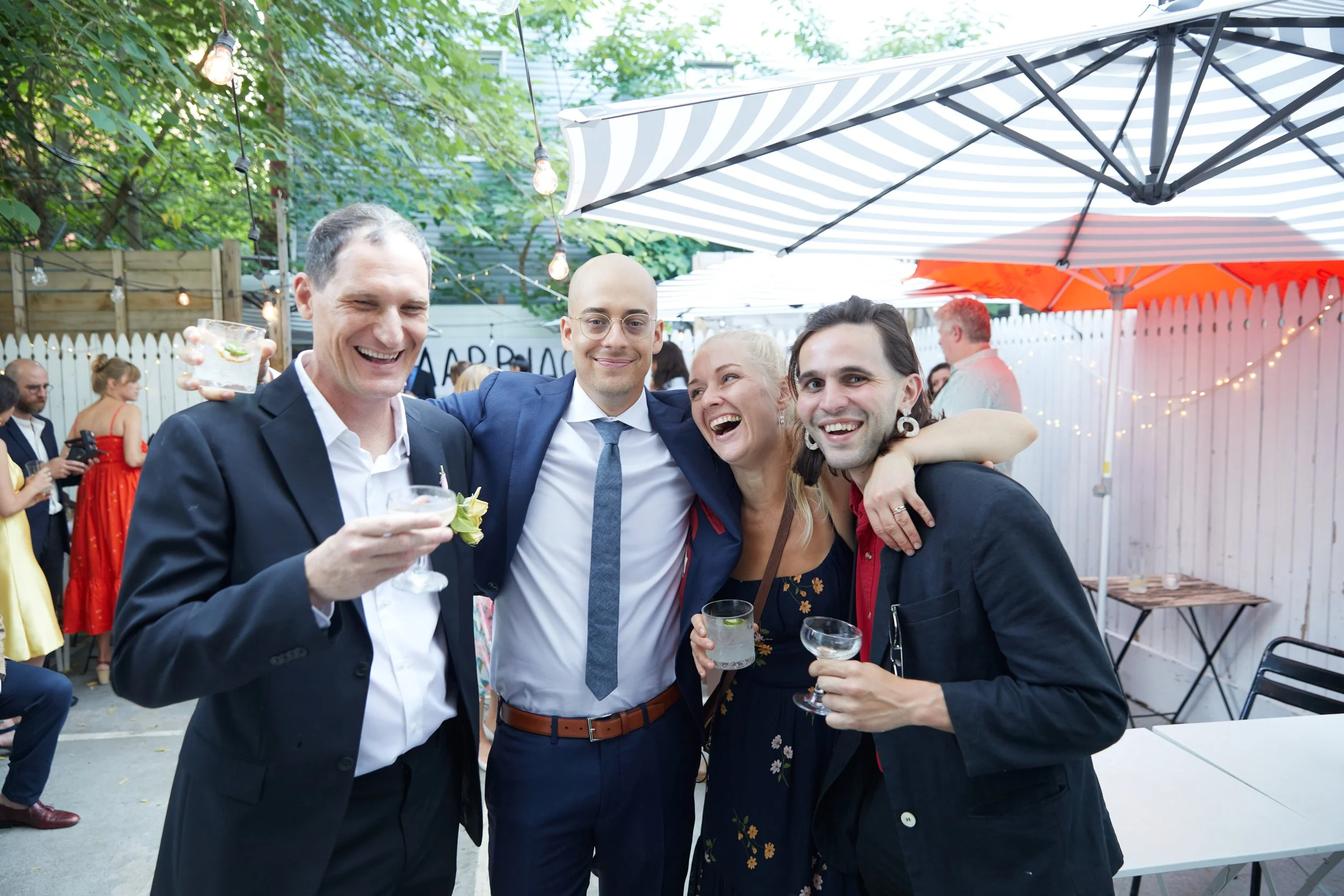 Group of four friends smiling and celebrating at an outdoor party, holding drinks, with string lights, umbrellas, and other guests in the background.