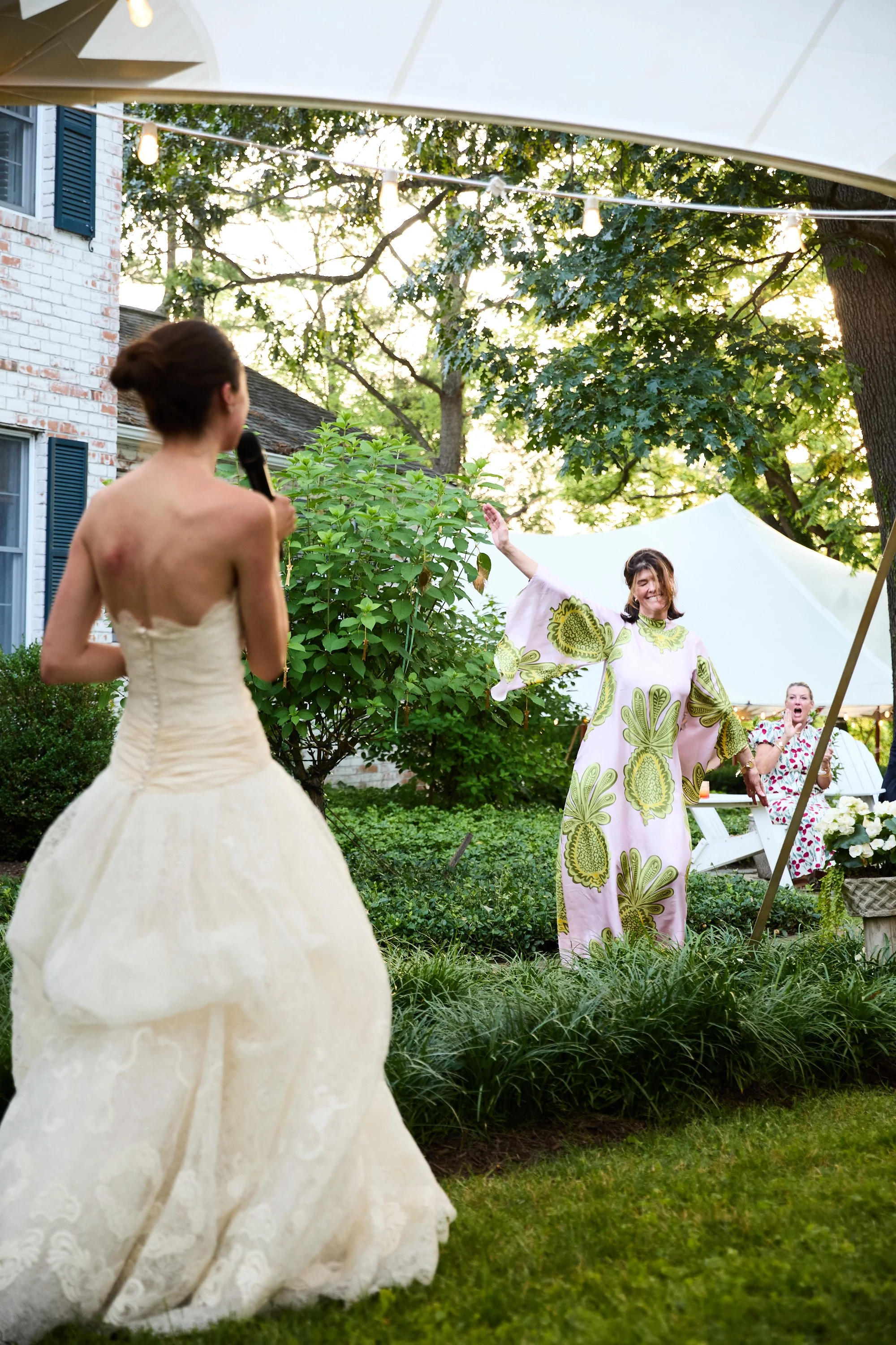 A woman in a wedding dress singing into a microphone in a garden with guests watching.