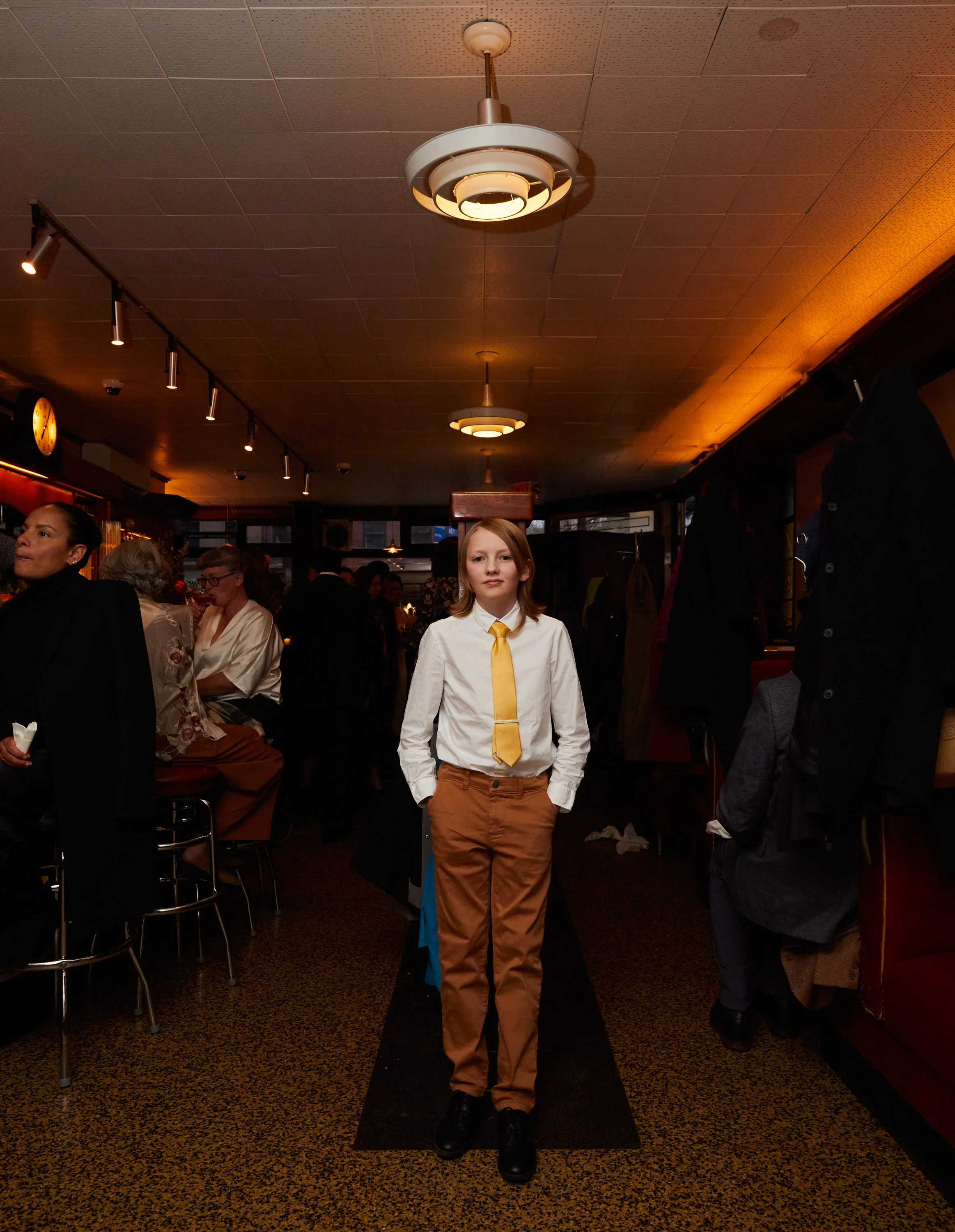 Young woman with red hair standing in the middle of a restaurant or bar, wearing a white shirt and yellow tie, with hands in her pockets, surrounded by other patrons.