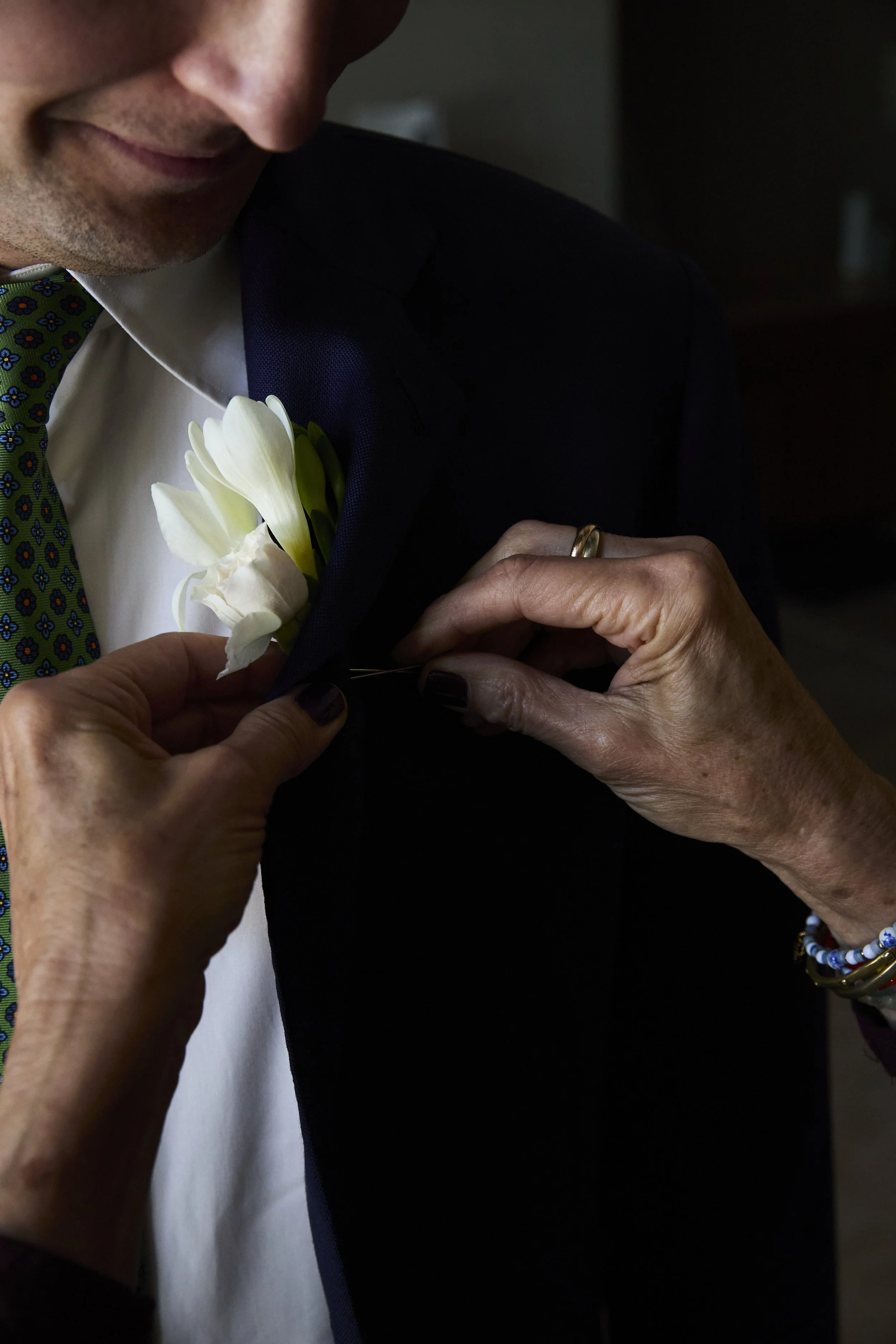 Close-up of a person pinning a white boutonniere and bouton on a man's dark suit.