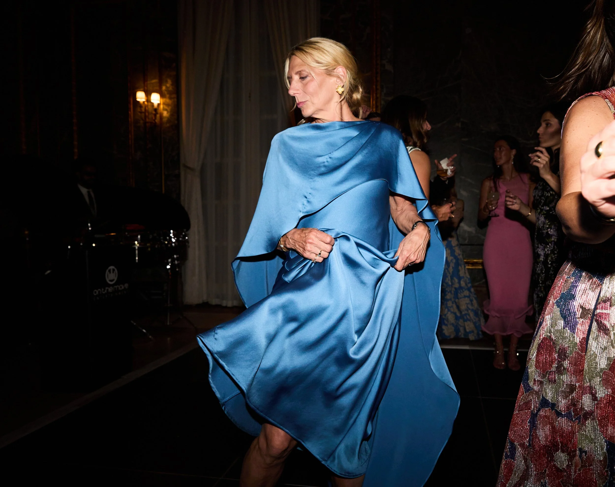 An older woman in a flowing, shiny blue dress dancing at a social event, with other women and a pianist in the background.