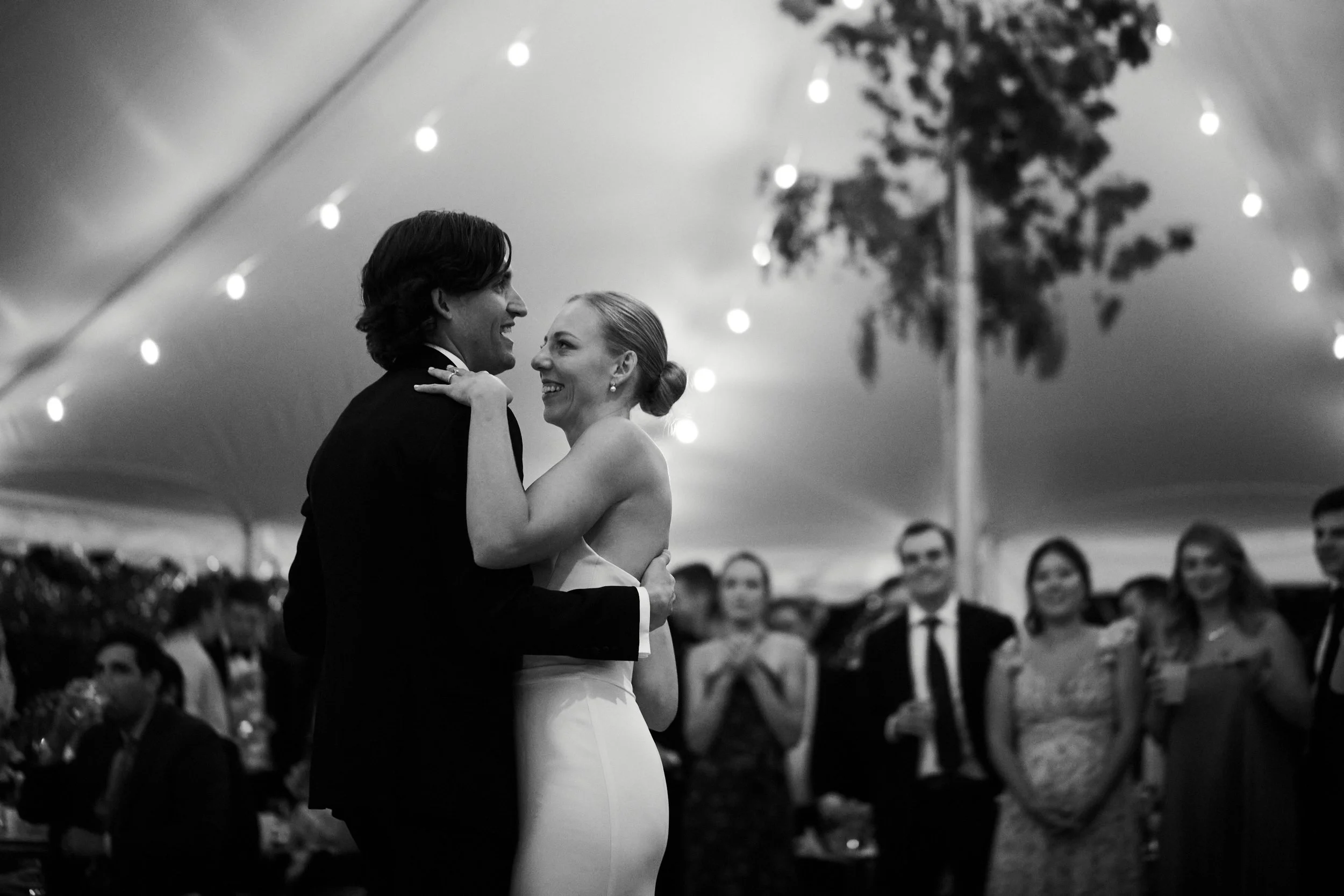 A bride and groom dance closely together at their wedding reception, smiling and gazing into each other's eyes, with wedding guests watching in the background under a decorated tent.