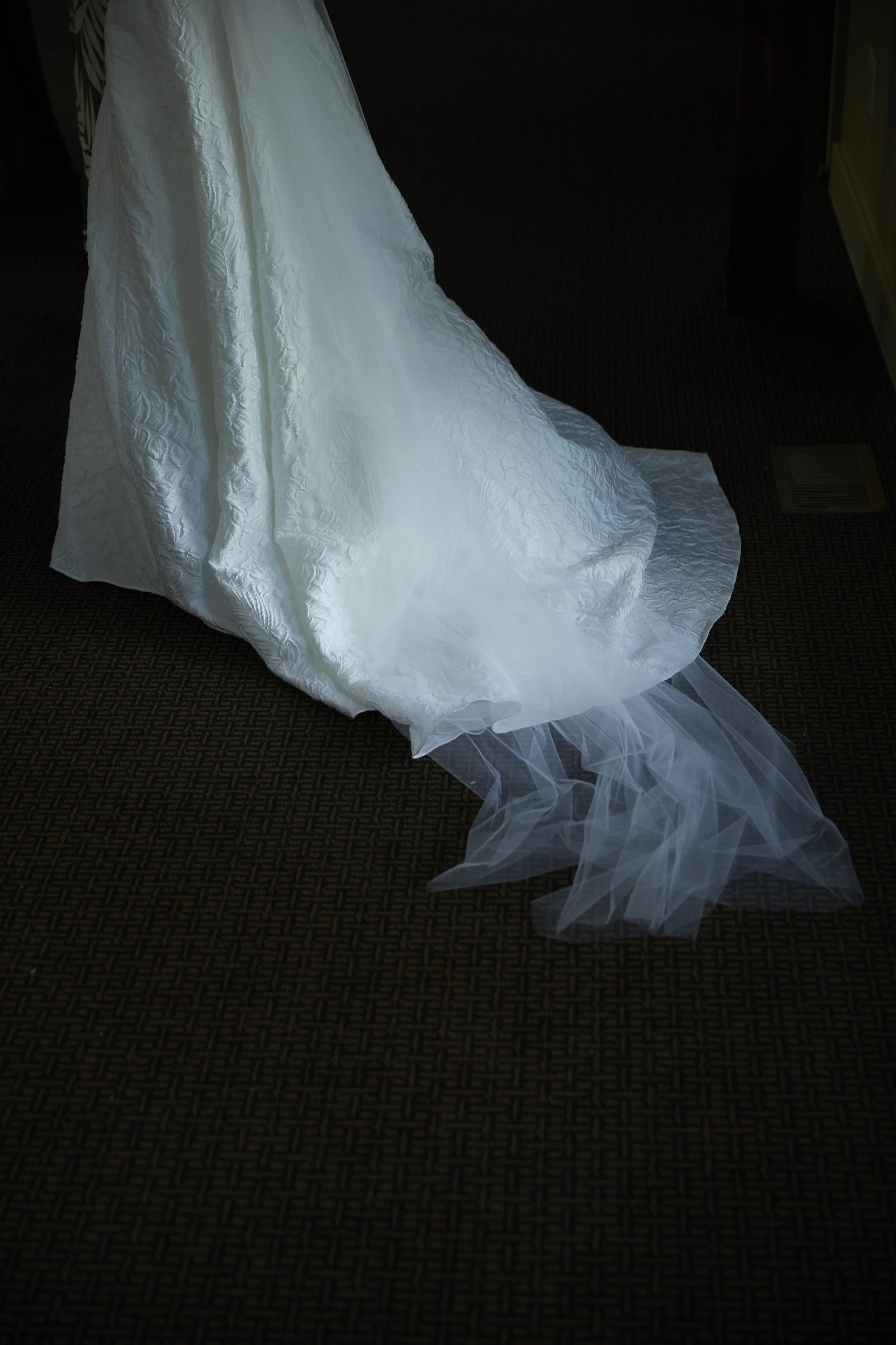 Close-up of a white wedding dress with a long train and a veil on a patterned carpet.