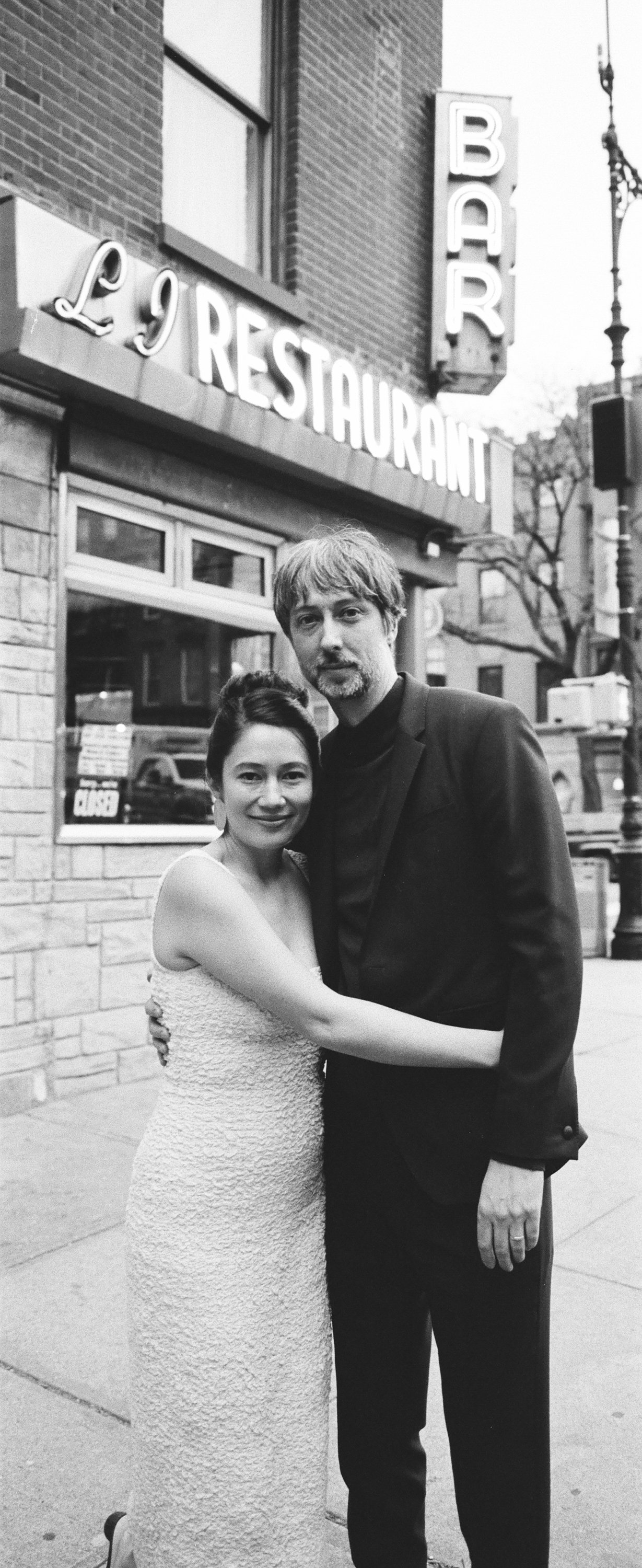 A black and white photo of a woman and a man standing on a city sidewalk, embracing each other in front of a restaurant with a neon sign that reads 'L9 RESTAURANT'. The woman is wearing a sleeveless dress, and the man is dressed in a dark suit.