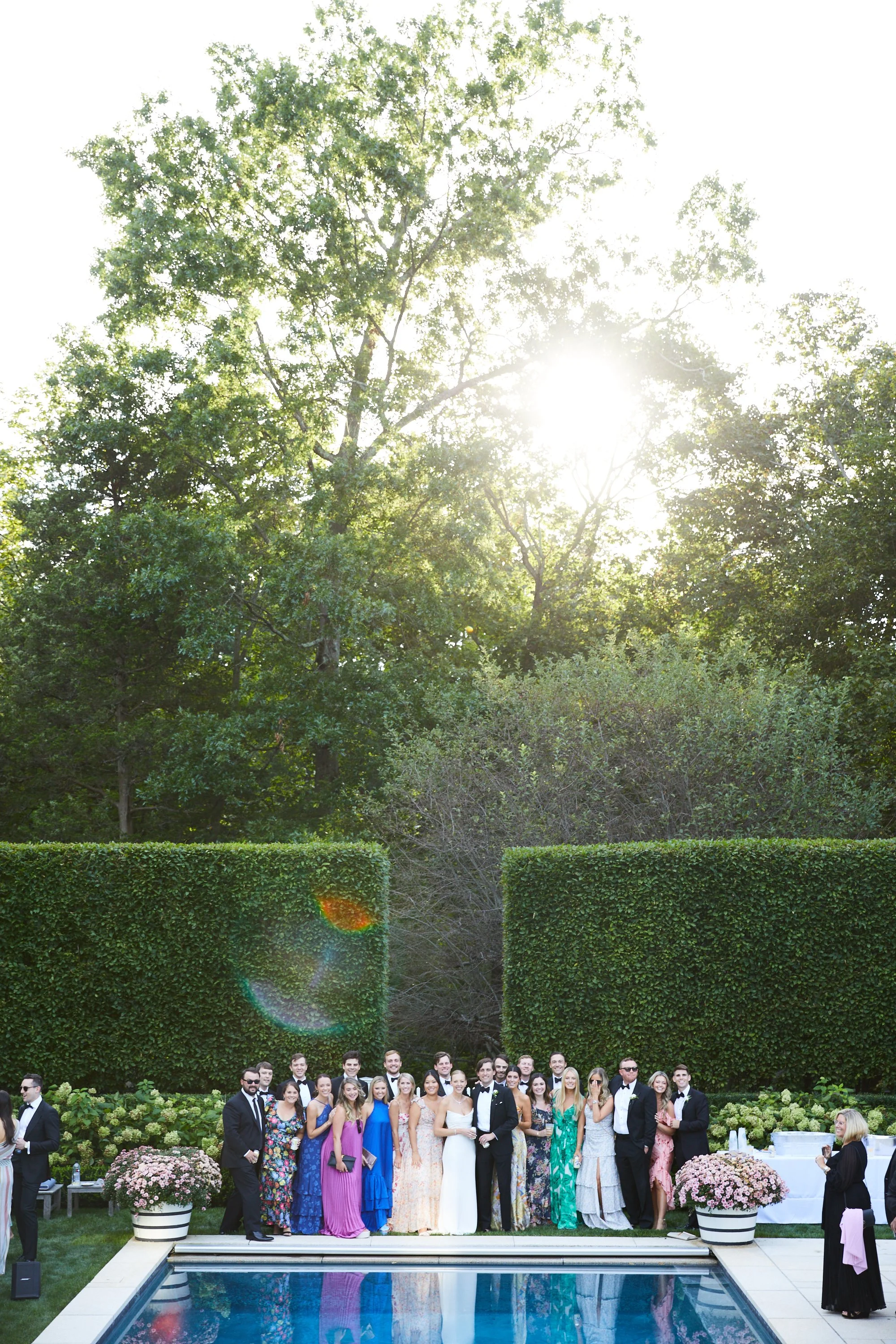 Group of people in formal attire standing by a pool at an outdoor event, surrounded by lush greenery and large hedges, with sunlight filtering through trees in the background.