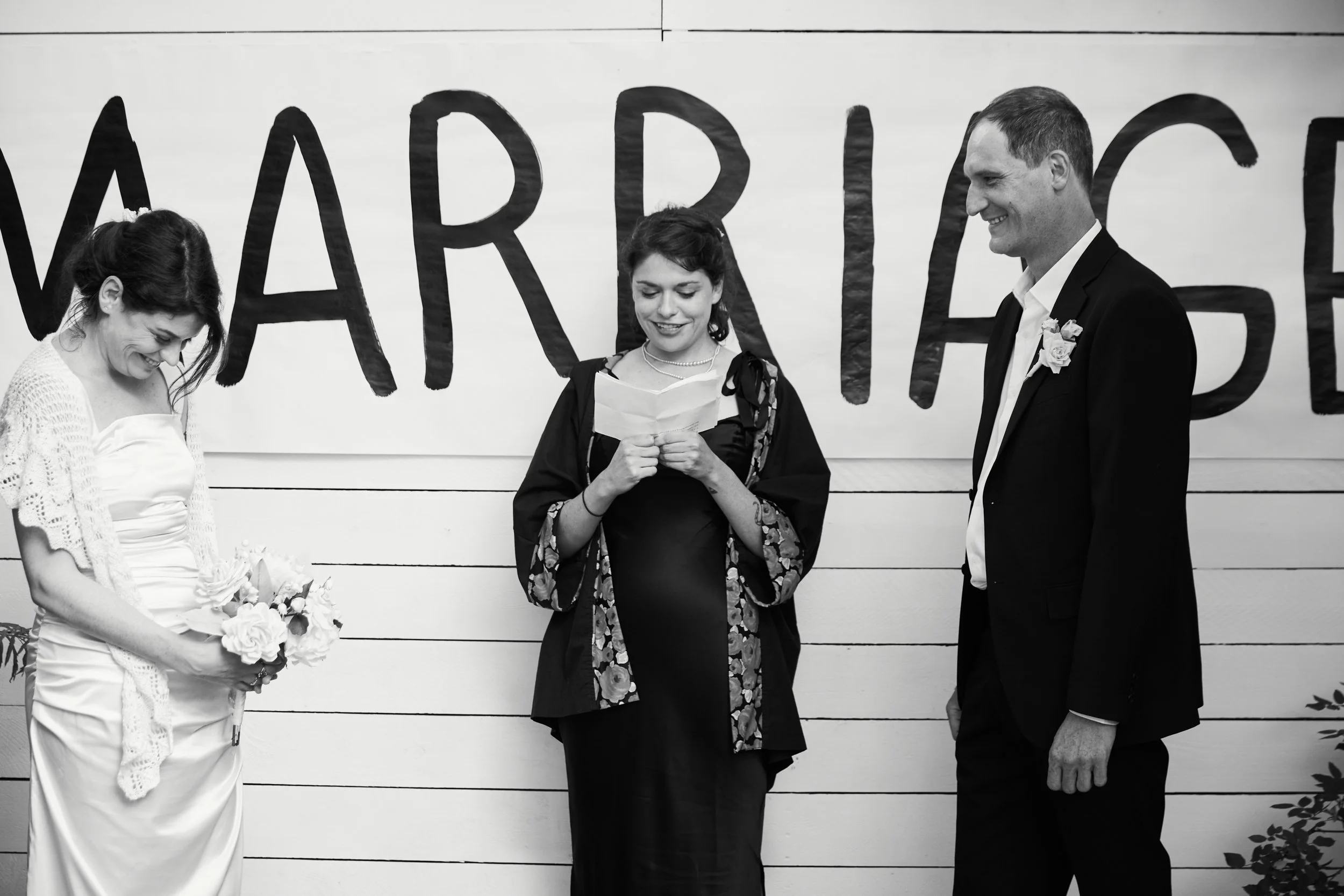 Three people at a wedding ceremony standing in front of a decorated wall with a large sign that spells out "Marriage." The woman on the left is the bride, holding a bouquet and looking down, smiling. The woman in the middle is reading from a piece of