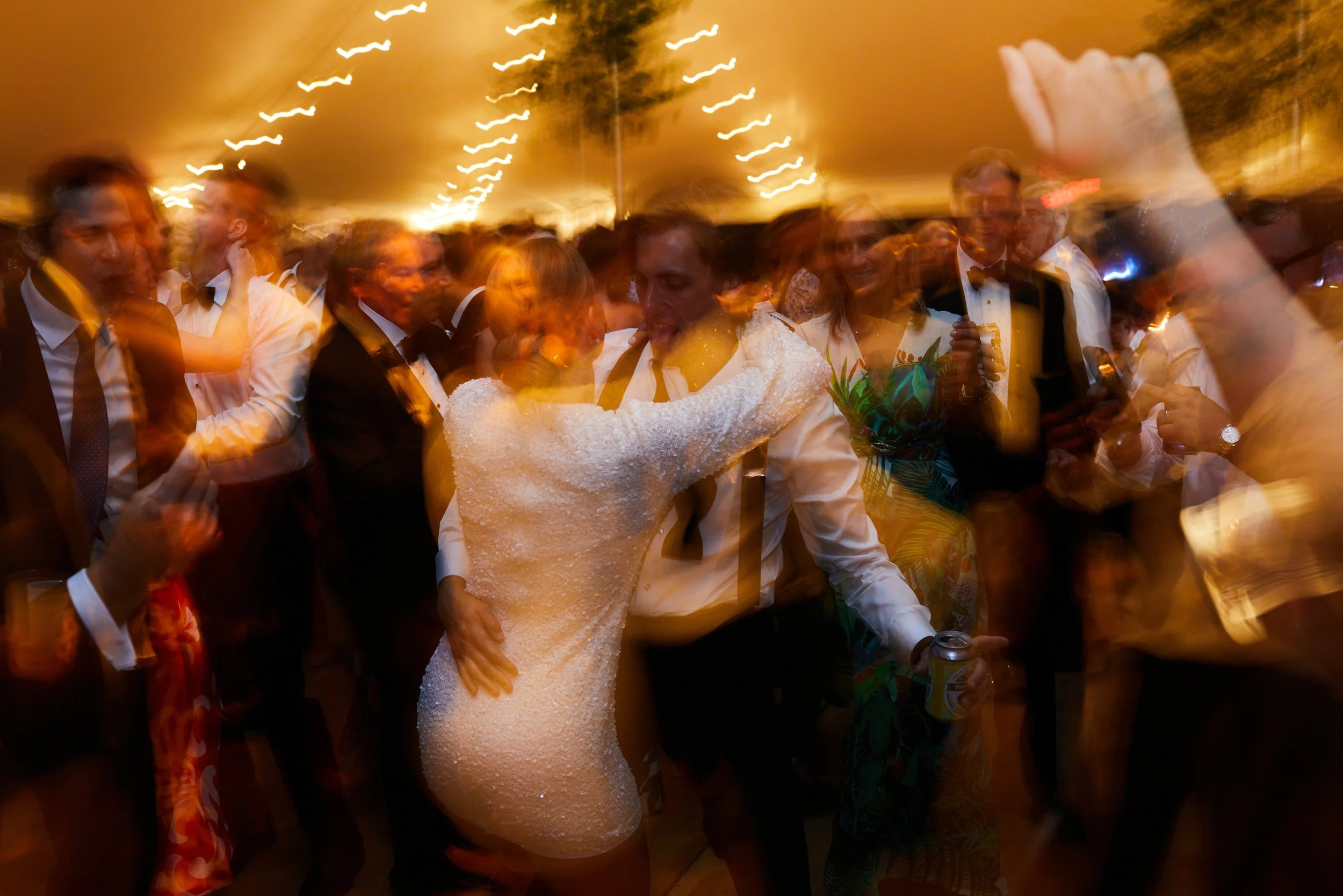 People dancing and celebrating at a lively party, with a woman in a white dress and a man holding a drink, surrounded by other guests in formal attire.