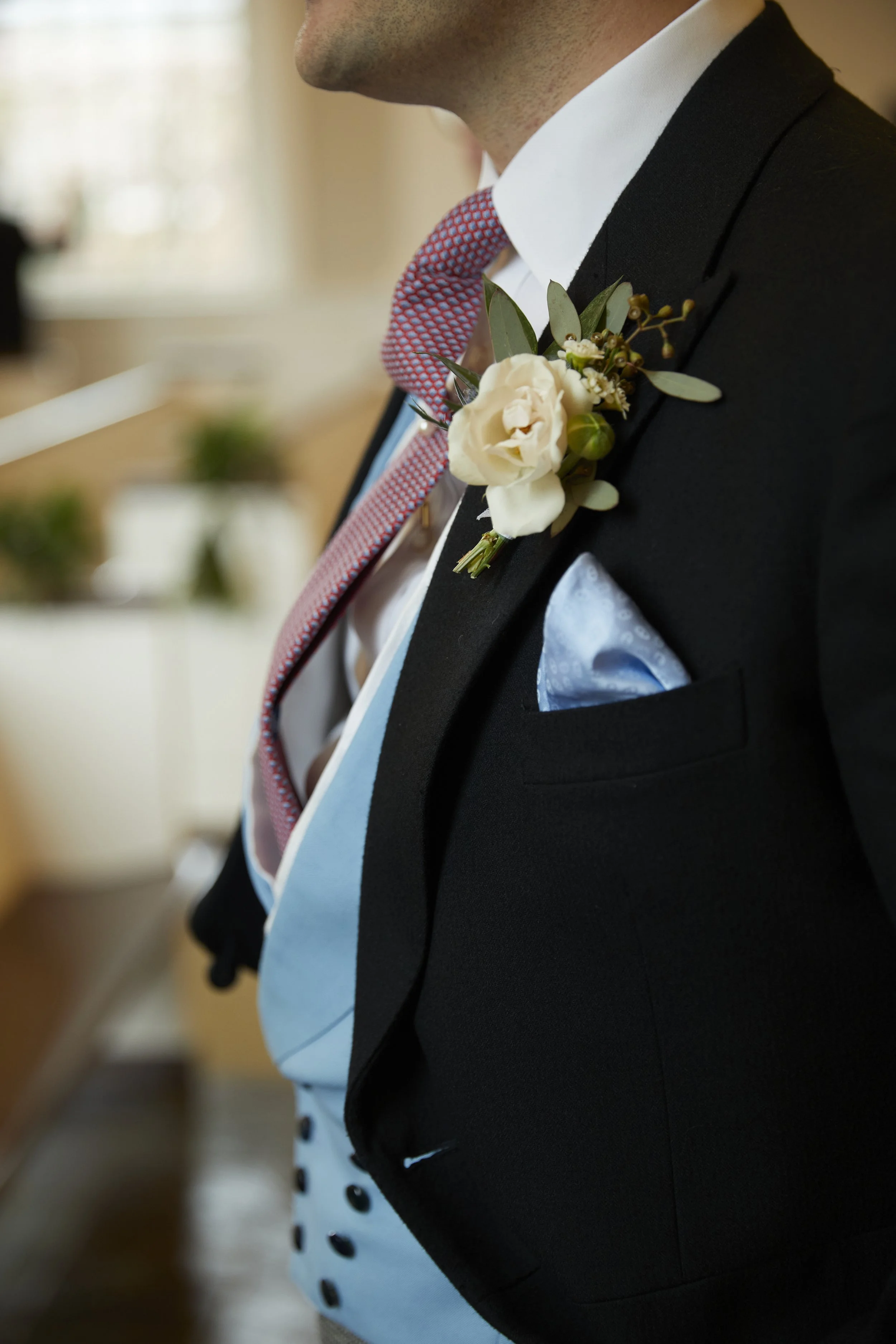 Close-up of a man dressed in formal wedding attire, wearing a black tuxedo with a sublimate boutonniere on his lapel, a white dress shirt, a pink and blue patterned tie, and a light blue vest with black buttons. A light blue pocket square is visible 