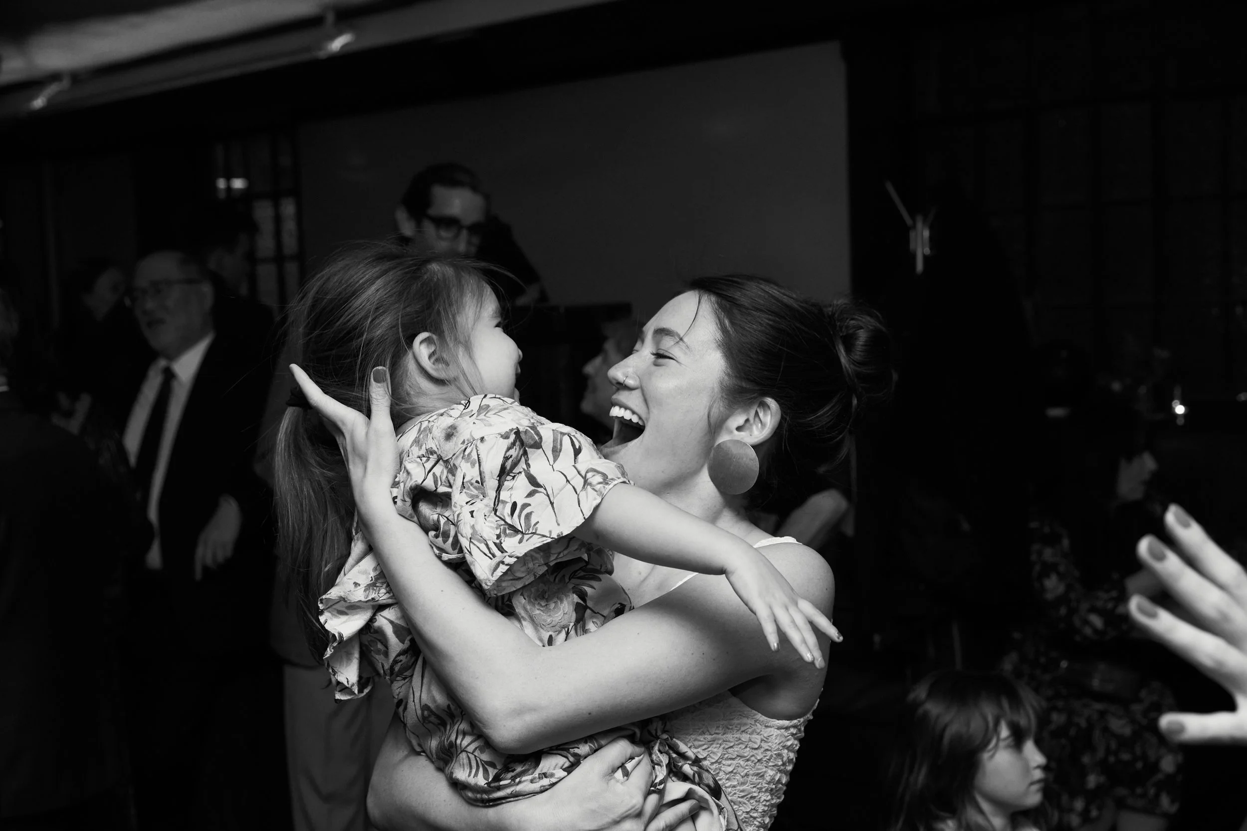 A woman with earrings is joyfully holding a girl in her arms, both smiling and touching faces. They are at a lively indoor event with other people in the background.