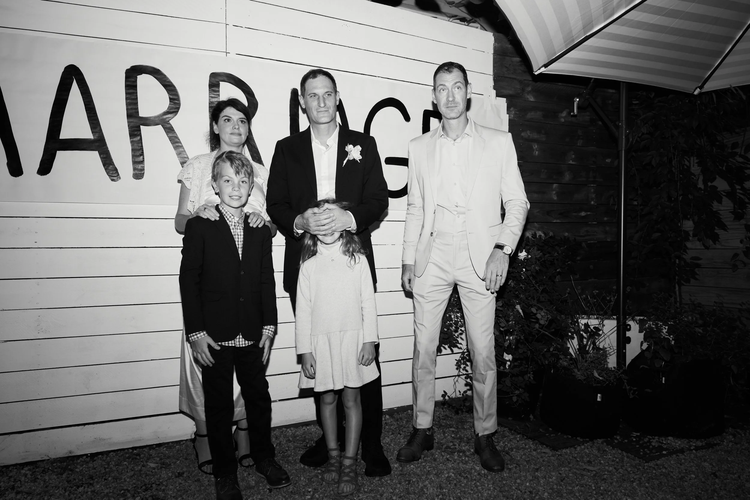 A group of five people, including two men, two women, and a boy, standing outdoors at night in front of a wall with large letters that spell out part of a word, possibly "MARRIAGE". They are dressed formally, with the men in suits and the women in dr