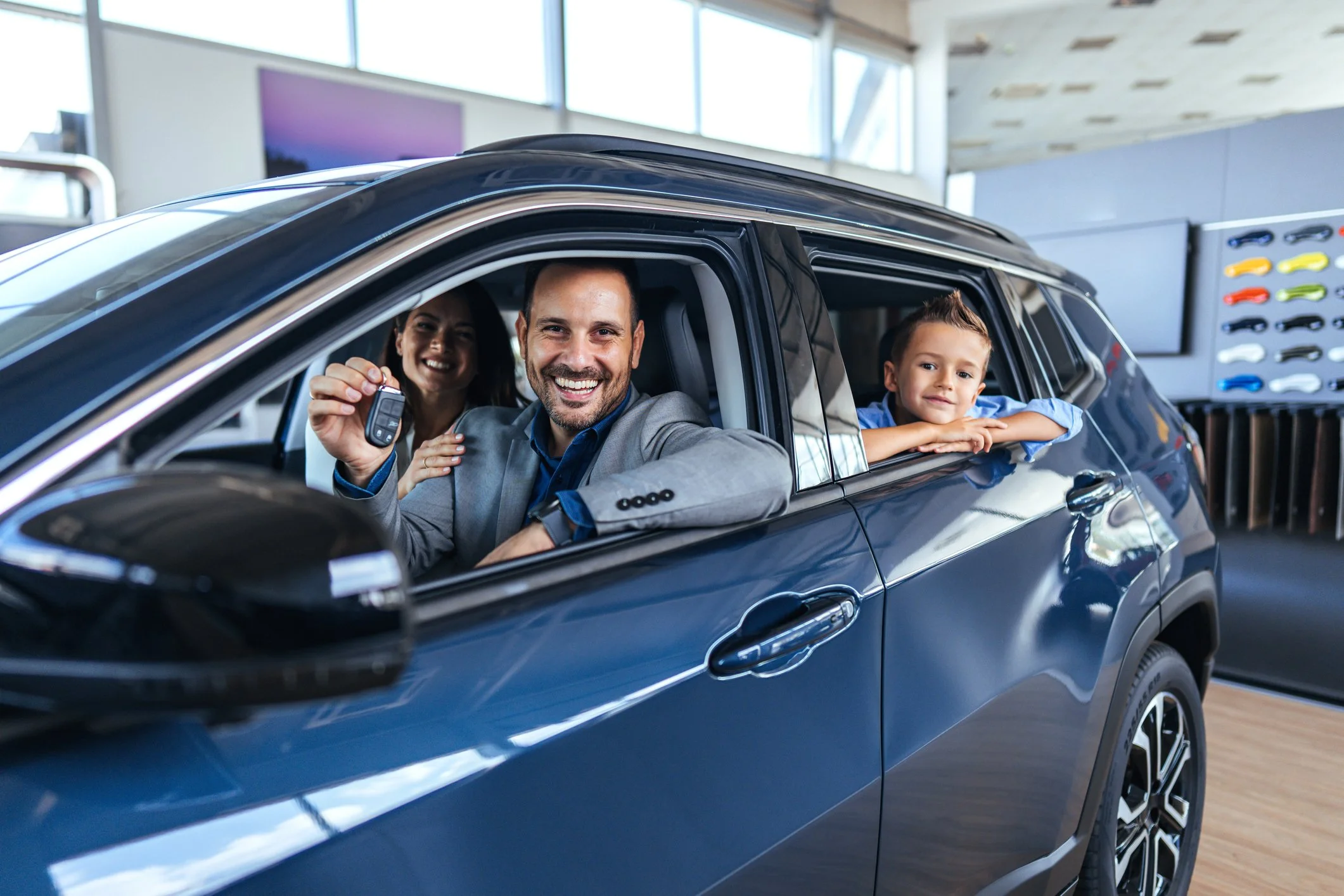 A happy man holding car keys inside a new dark blue SUV at a car dealership, with a woman and a young boy also in the car.