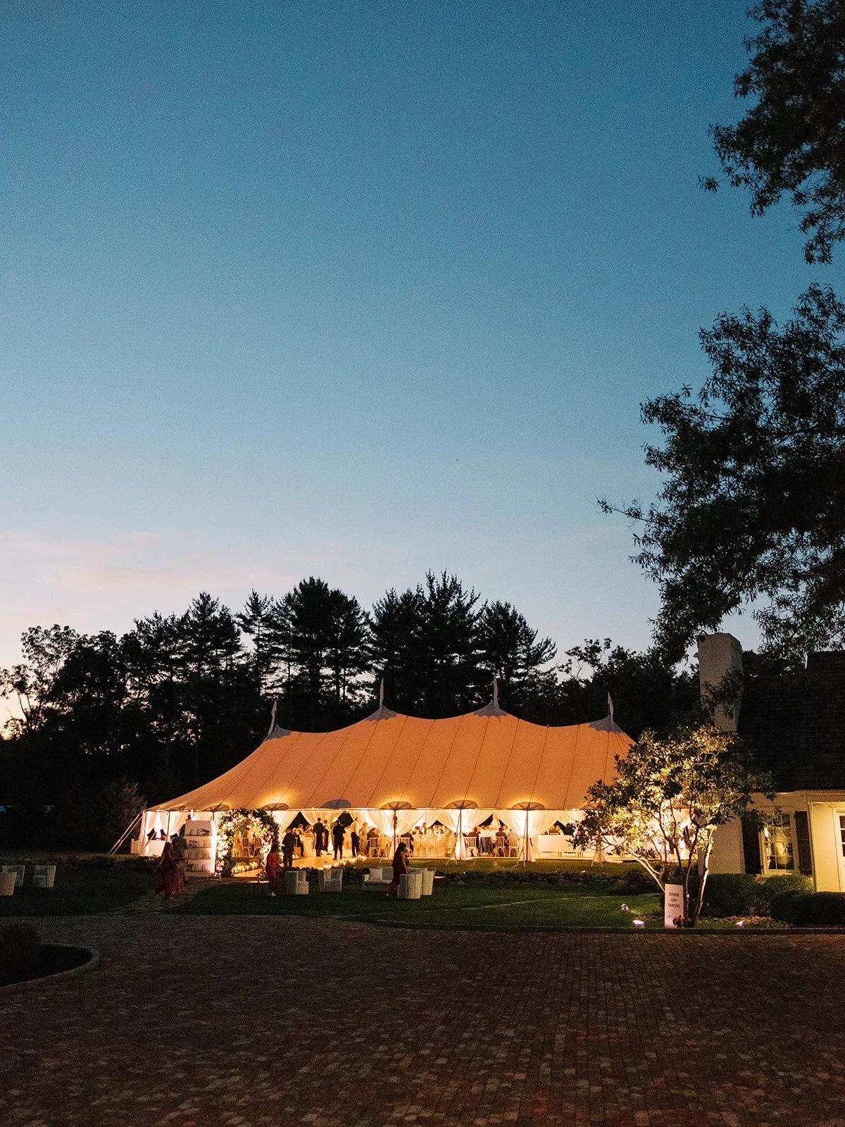 An outdoor wedding reception under a large, illuminated tent during dusk with people gathered inside and outside, surrounded by trees and a brick pathway.