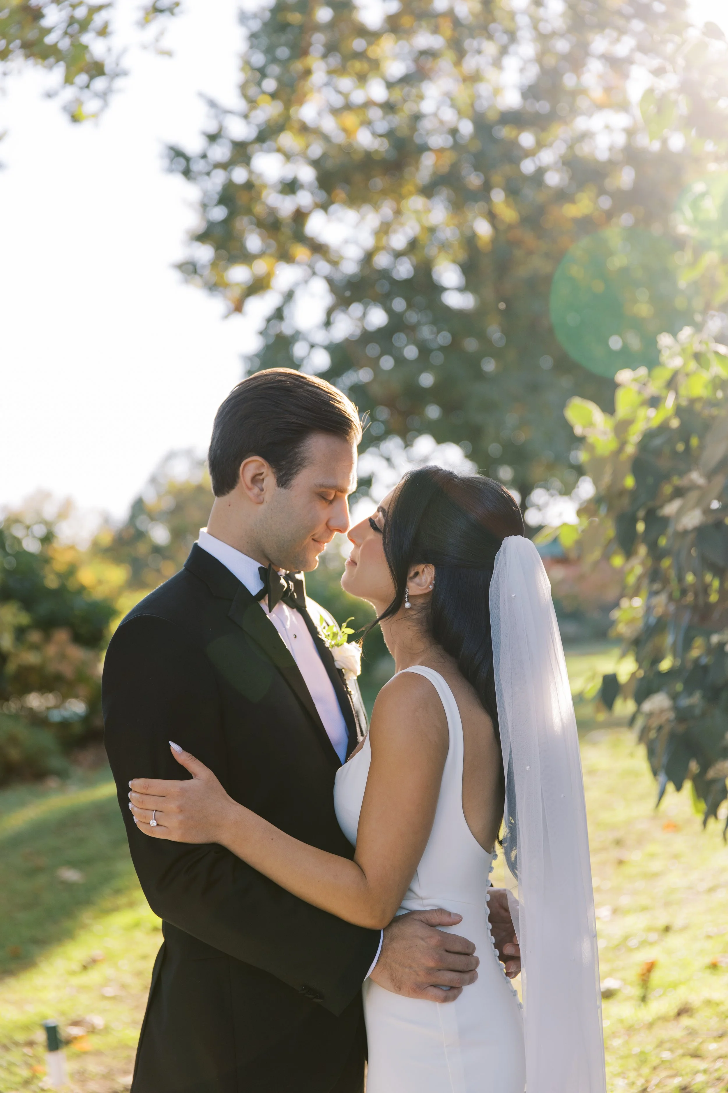 A bride and groom embrace outdoors, about to kiss, with trees and sunlight in the background.