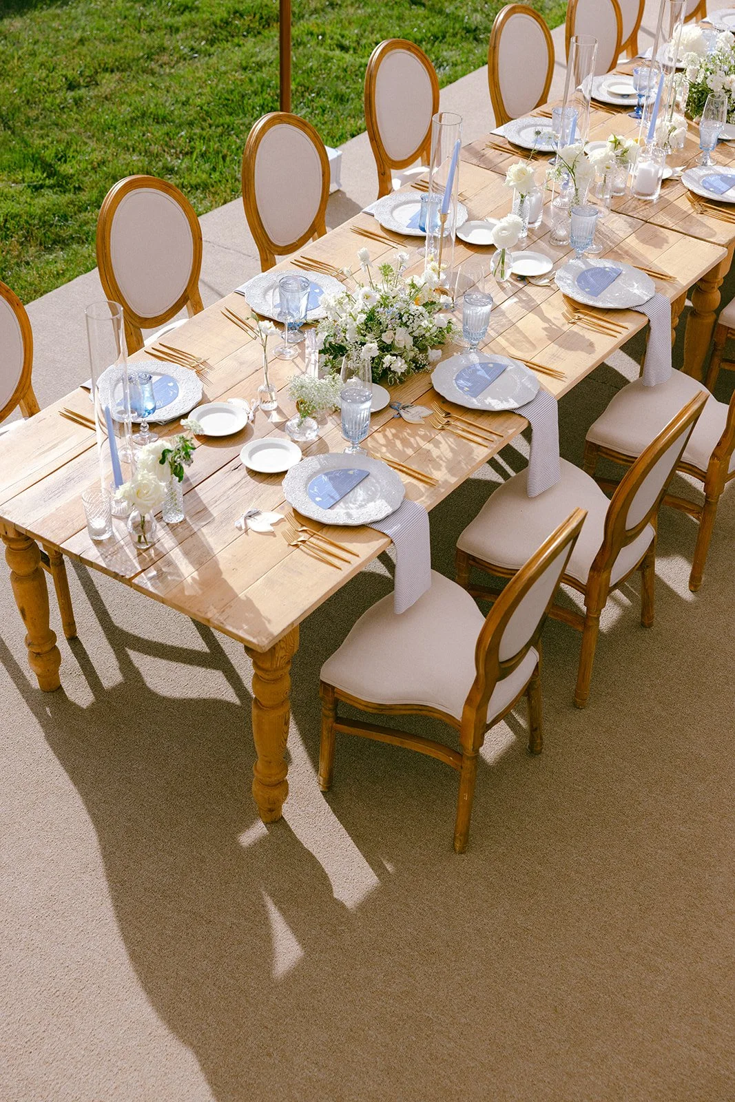 Outdoor dining table set for a formal event with plates, glasses, cutlery, floral centerpieces, and cloth napkins, surrounded by beige chairs with wooden frames.