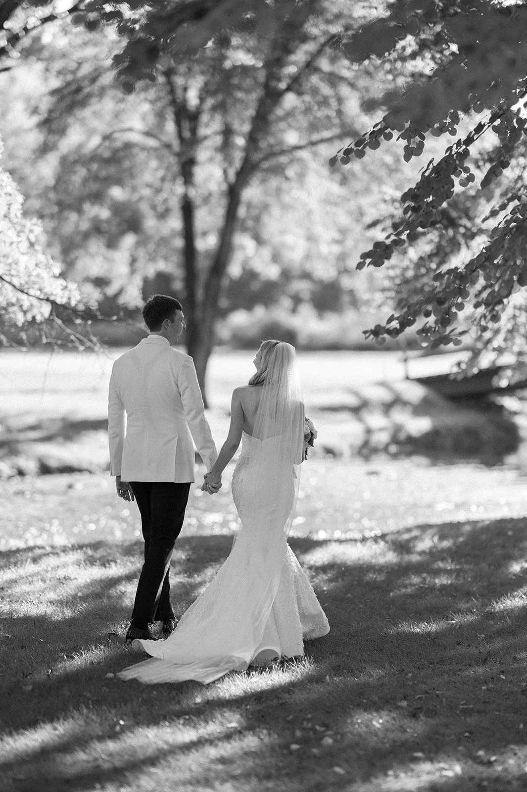 A black and white photo of a bride and groom walking hand in hand outdoors near a lake, surrounded by trees.