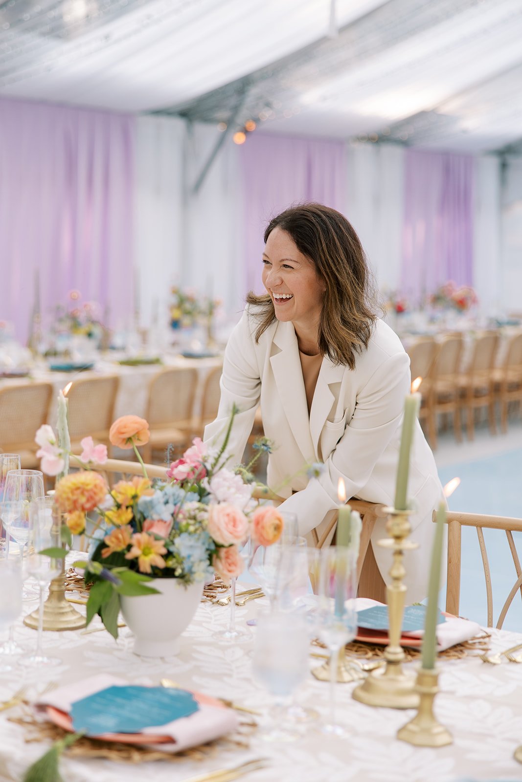 Woman smiling and arranging a flower centerpiece at a decorated event table, with pastel-colored flowers, candles, and table settings, in a brightly lit tent with purple curtains.