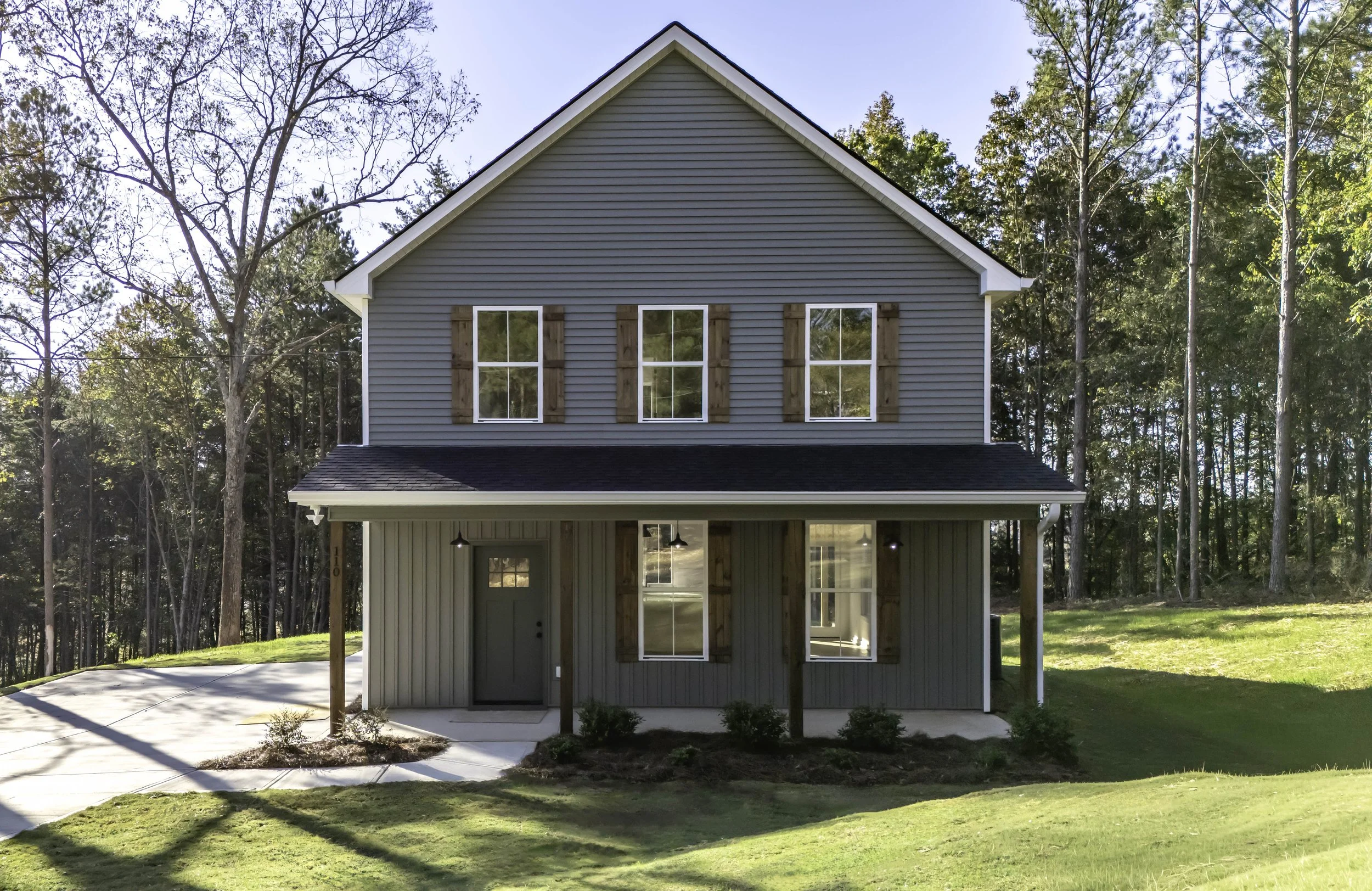 New two-story house with gray siding and brown window shutters, front porch with wooden posts, surrounded by green lawn and trees, in a rural setting.