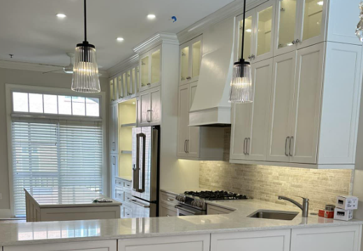 Elegant custom white shaker kitchen cabinets with floor-to-ceiling storage, illuminated glass display cabinets, & a custom range hood. Designed to maximize storage, functionality, and timeless style while showcasing expert craftmanship