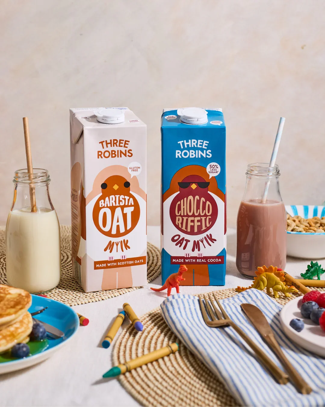 Two cartons of oat milk, one labeled "Barista Oat" and the other "Chocco Riffic Oat Milk," placed on a table with breakfast items, including a bowl of cereal, a plate of pancakes with blueberries and syrup, small dinosaur toys, and utensils on a striped cloth napkin. There are also two glass bottles with straws containing milk and chocolate milk.
