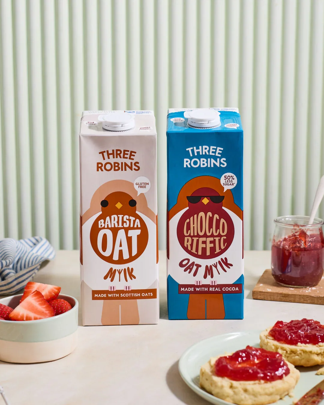 Two cartons of oat milk from Three Robins, one labeled Barista Oat and the other Chocco Riffic, placed on a table with strawberries, jam, and biscuits with strawberry jam in the background.