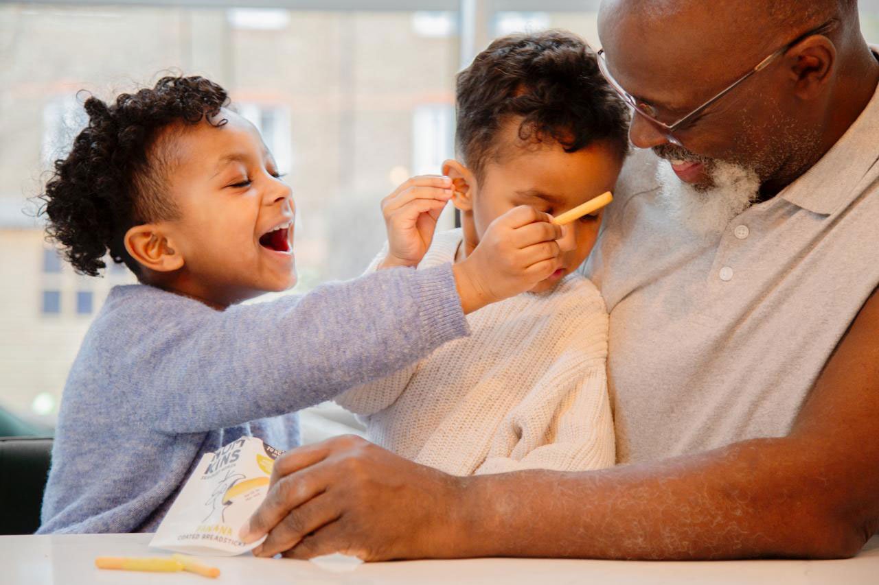 A young girl playfully putting a stick on her young brother's nose while their grandfather smiles and watches, sitting at a table.