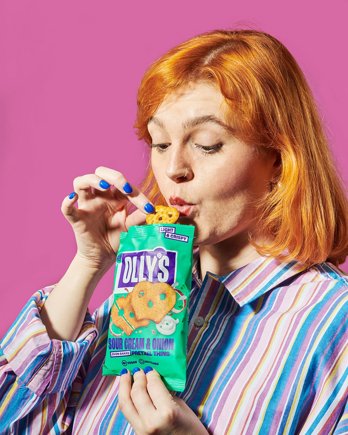Woman with red hair and blue nail polish tasting a sour cream and onion flavored snack from a bright green bag labeled Olly's against a pink background.