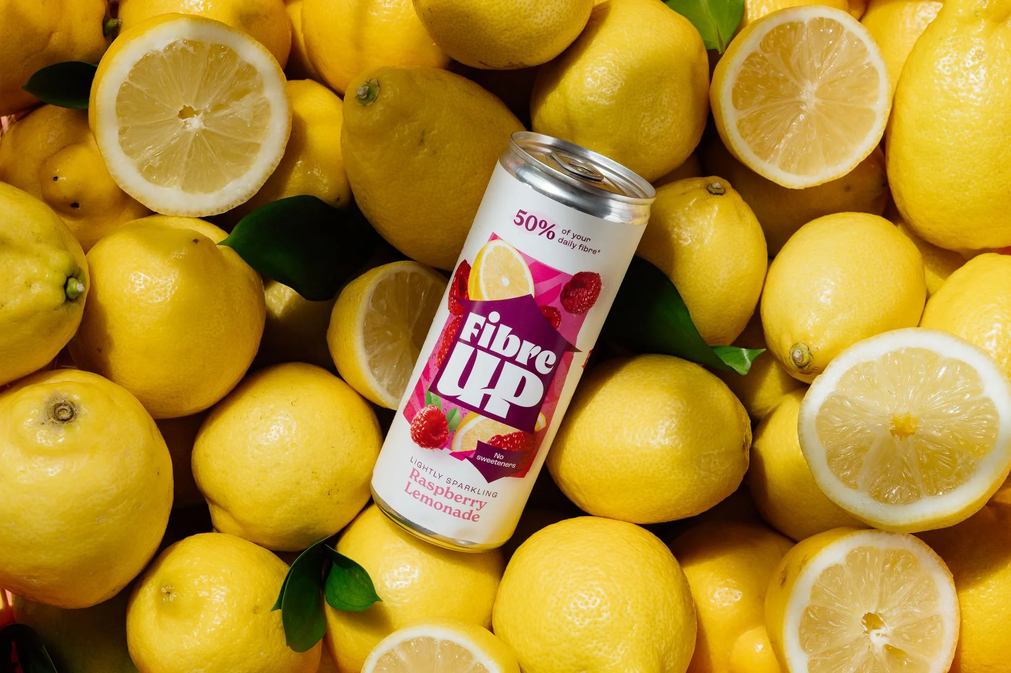 A can of Fibre UP Raspberry Lemonade sparkling drink resting among numerous fresh yellow lemons, some sliced in half, with green leaves scattered around.