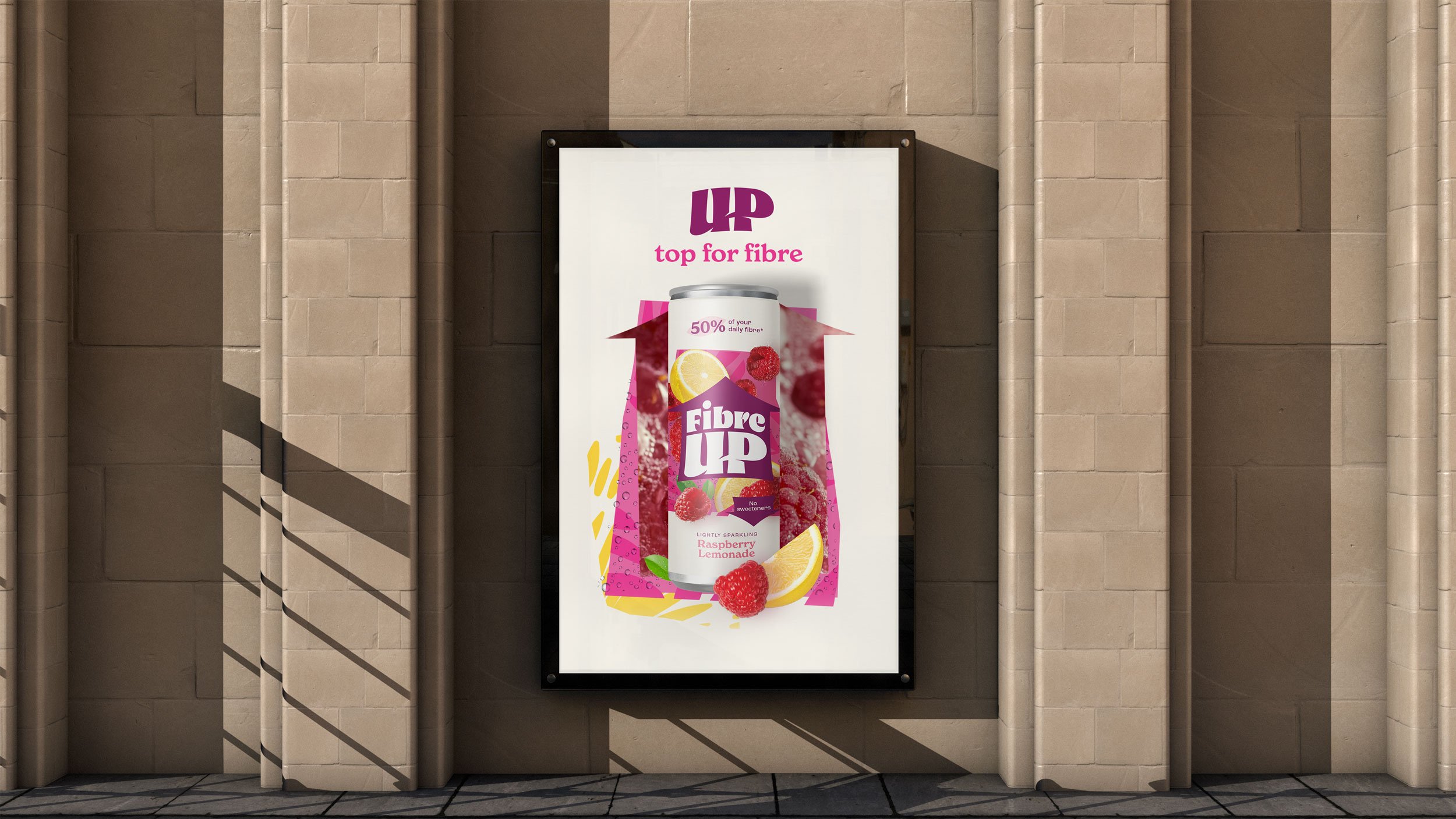 Poster on a beige brick wall advertising Fibre UP raspberry lemonade drink. The poster features an illustration of a can of Fibre UP with lemon and raspberry images, pink and yellow abstract shapes, and text highlighting the flavor and health benefits.