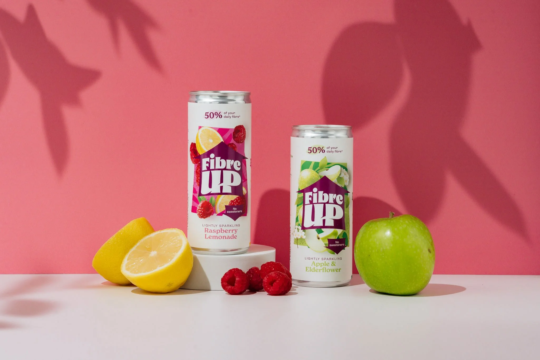 Two cans of Fibre Up lightly sparkling fruit-flavored drinks, one raspberry lemonade and one apple and elderflower, placed on a white surface with lemons, raspberries, and an apple, against a pink background with shadowed leaf shapes.