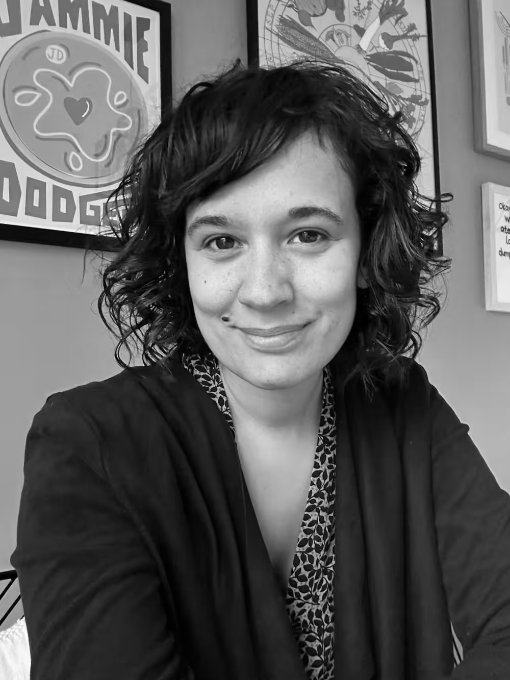 A woman with curly hair and a patterned shirt, smiling at the camera, sitting indoors with framed artwork on the wall behind her.