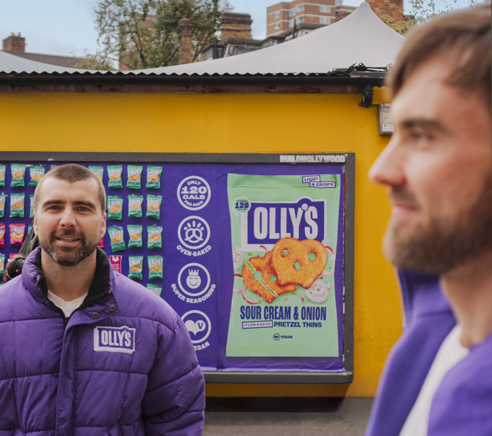 Two men standing in front of a bright yellow booth with a purple Olly's snack display behind them. One man wears a purple jacket with an Olly's logo, and the other is in a purple hoodie. A poster advertises Olly's sour cream and onion pretzel thins, highlighting features like low calories, oven-baked, super seasoning, and vegan.