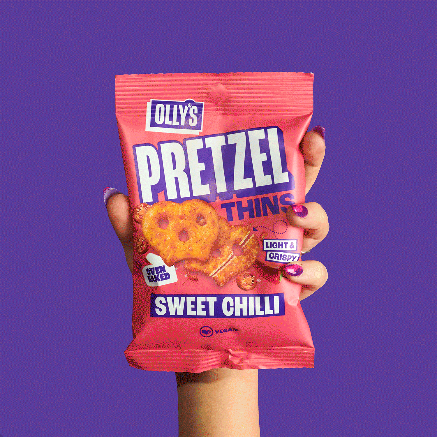 Hand holding a pink bag of Olly's Pretzel Thins, Sweet Chilli flavor, with images of pretzels on the packaging, against a purple background.