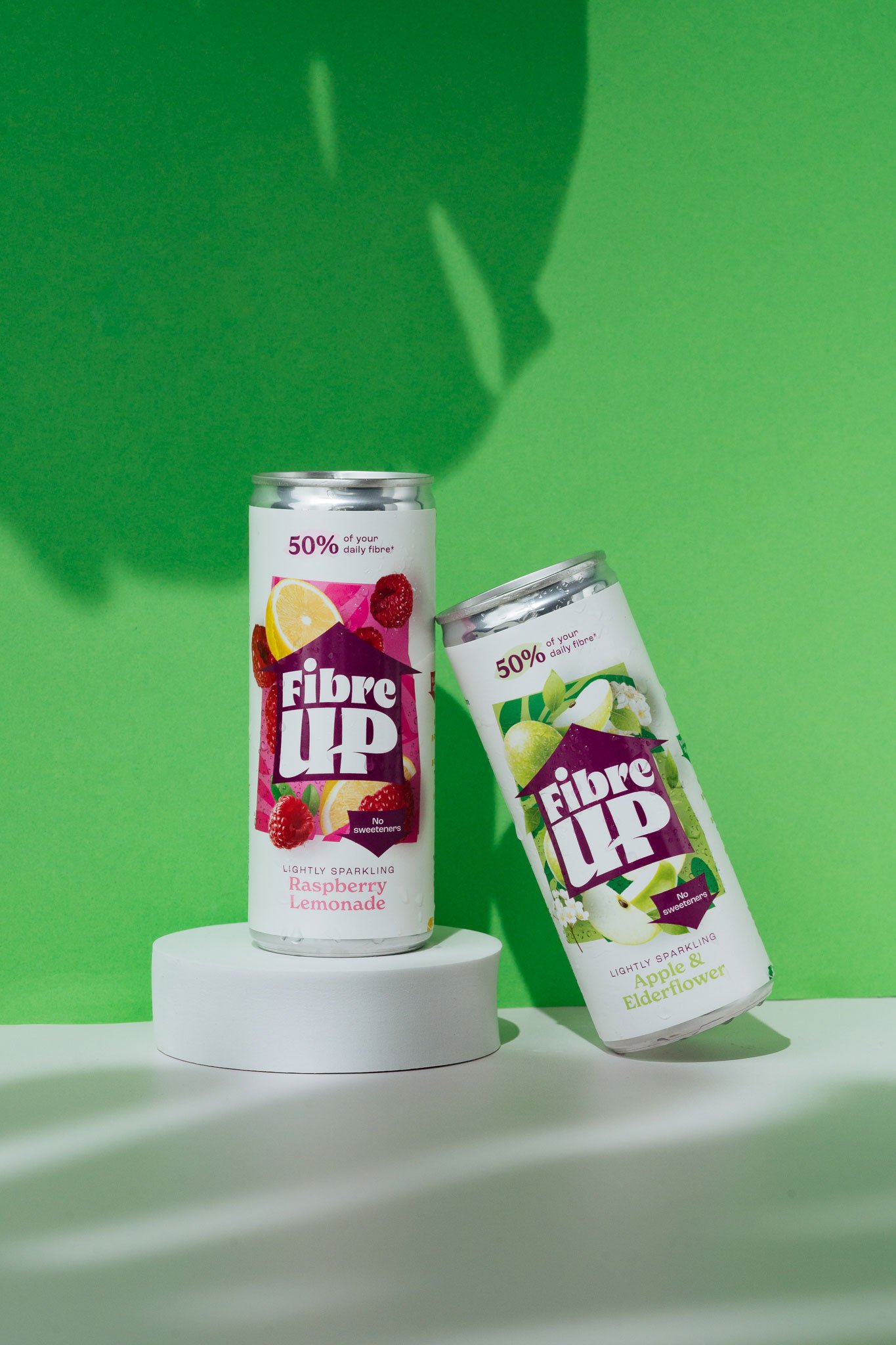 Two cans of Fibre UP lightly sparkling beverages, one raspberry lemonade and one apple elderflower, placed on white surfaces against a green background.
