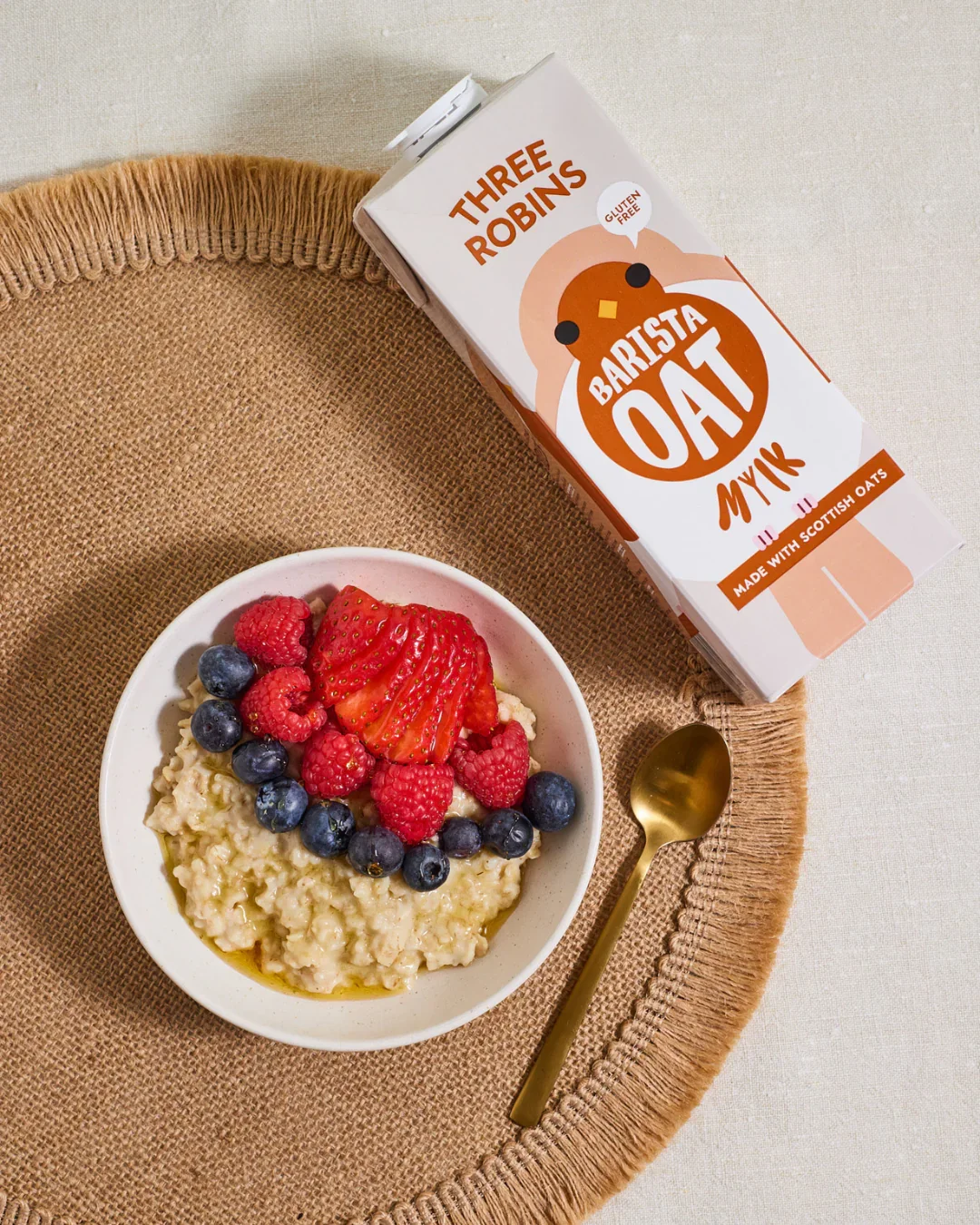 A bowl of oatmeal topped with sliced strawberries, raspberries, and blueberries on a round, woven placemat. Next to the bowl is a gold spoon, and a carton of Three Robins Barista Oat Milk placed nearby.