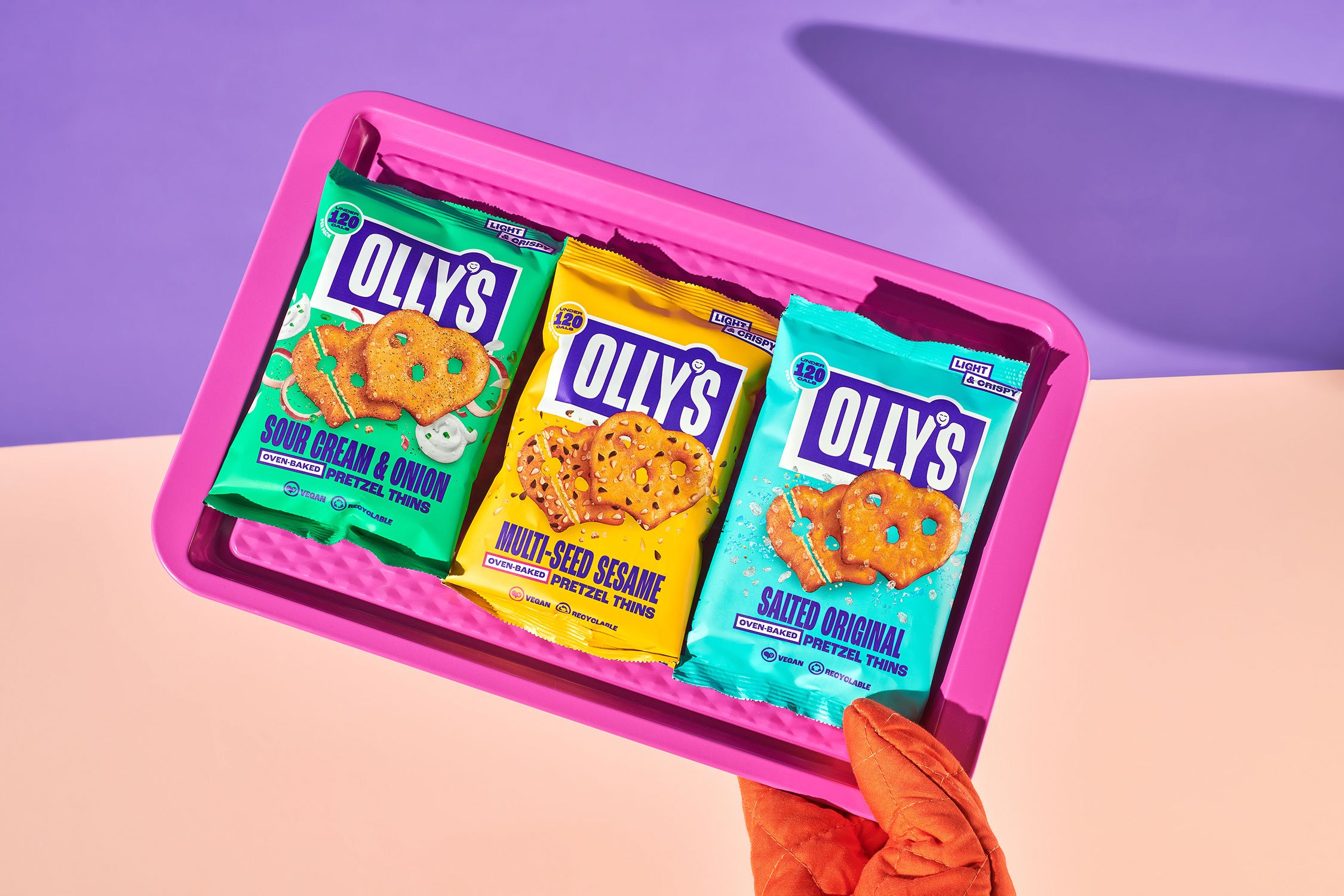 A pink tray holding three bags of Olly's pretzel thins in different flavors: sour cream & onion, multi-seed sesame, and salted original. A gloved hand is holding the tray.