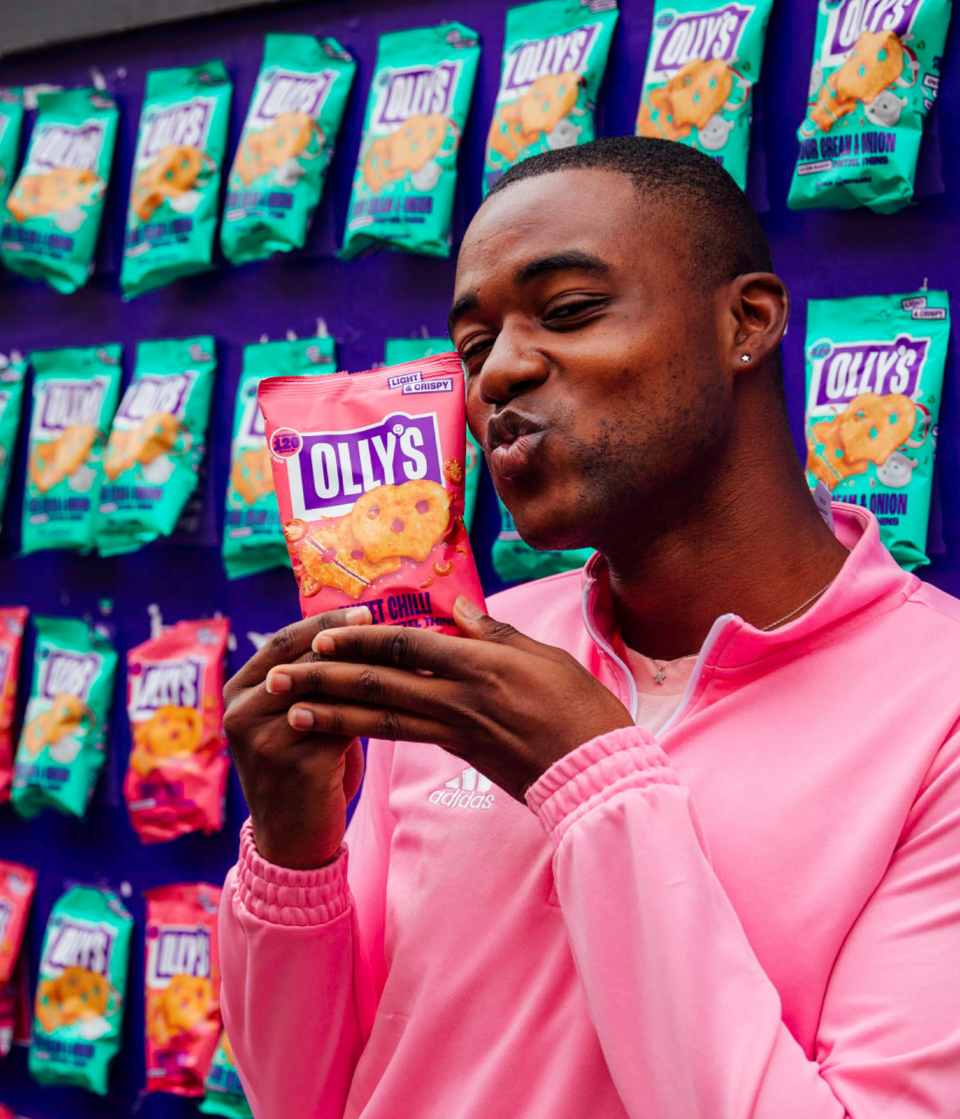 A man in a pink Adidas jacket holding a bag of Olly's potato chips, with many more chip bags displayed on a wall behind him.