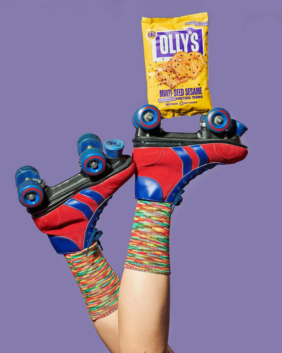 Close-up of a person's legs and feet wearing colorful socks and red and blue roller skates, holding a bag of Olly's multi-seed sesame pretzel thins on a purple background.