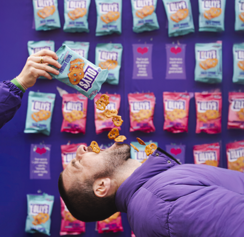 A person in a purple jacket is leaning back with their mouth open as another person, wearing a purple jacket, holds a bag of Lays chips and is pouring some into their mouth. The background shows a wall with purple, blue, and pink packets of Lays chips hanging.