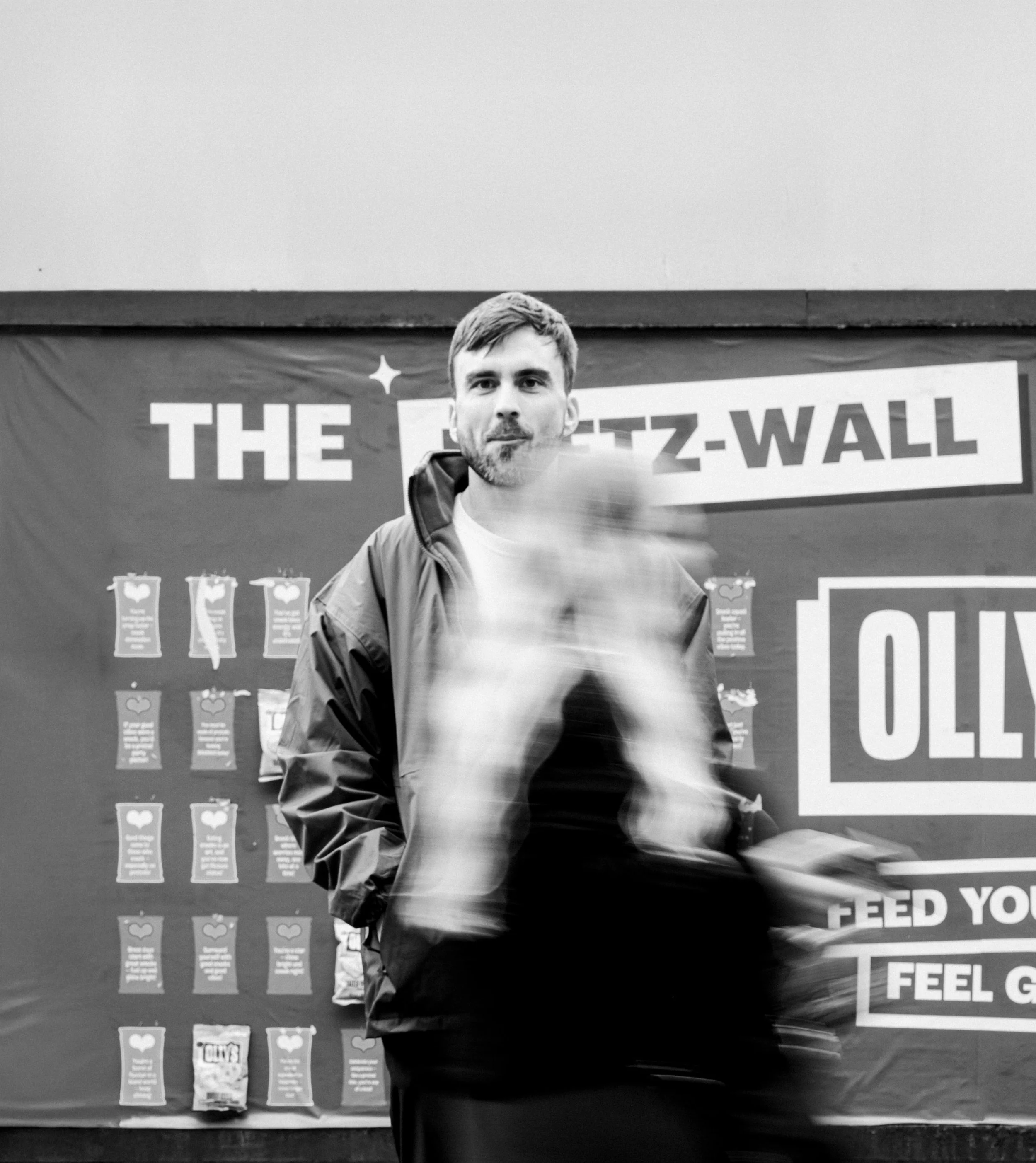 A man standing in front of a poster with the text 'THE Z- WALL' in large letters, with a blurred person walking past in the foreground.