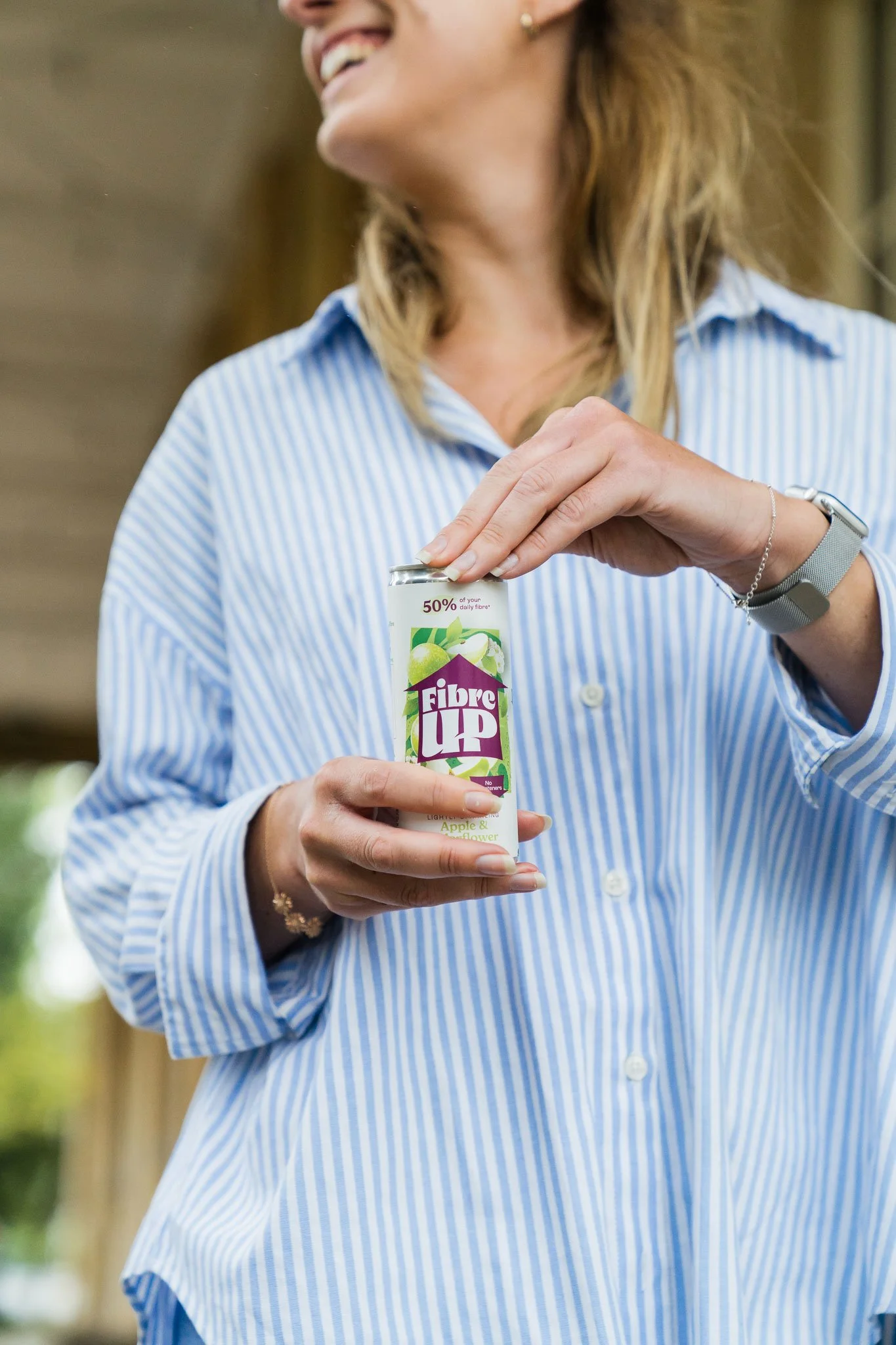 Woman holding a can of Fibre Up apple and flower flavored dietary supplement drink outdoors, smiling.