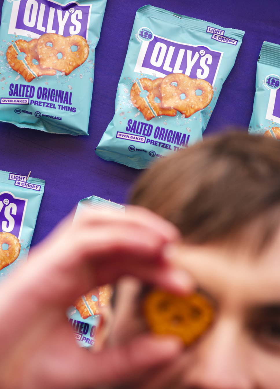 Multiple bags of Olly's Salted Original Pretzel Thins on a purple surface, with a blurred child holding a pretzel in front of the camera.