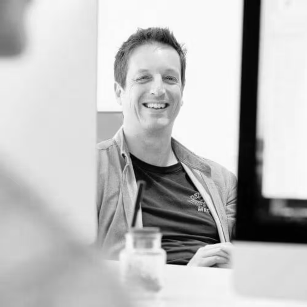 Smiling man in an office sitting at a desk, viewed partially through computer monitors.