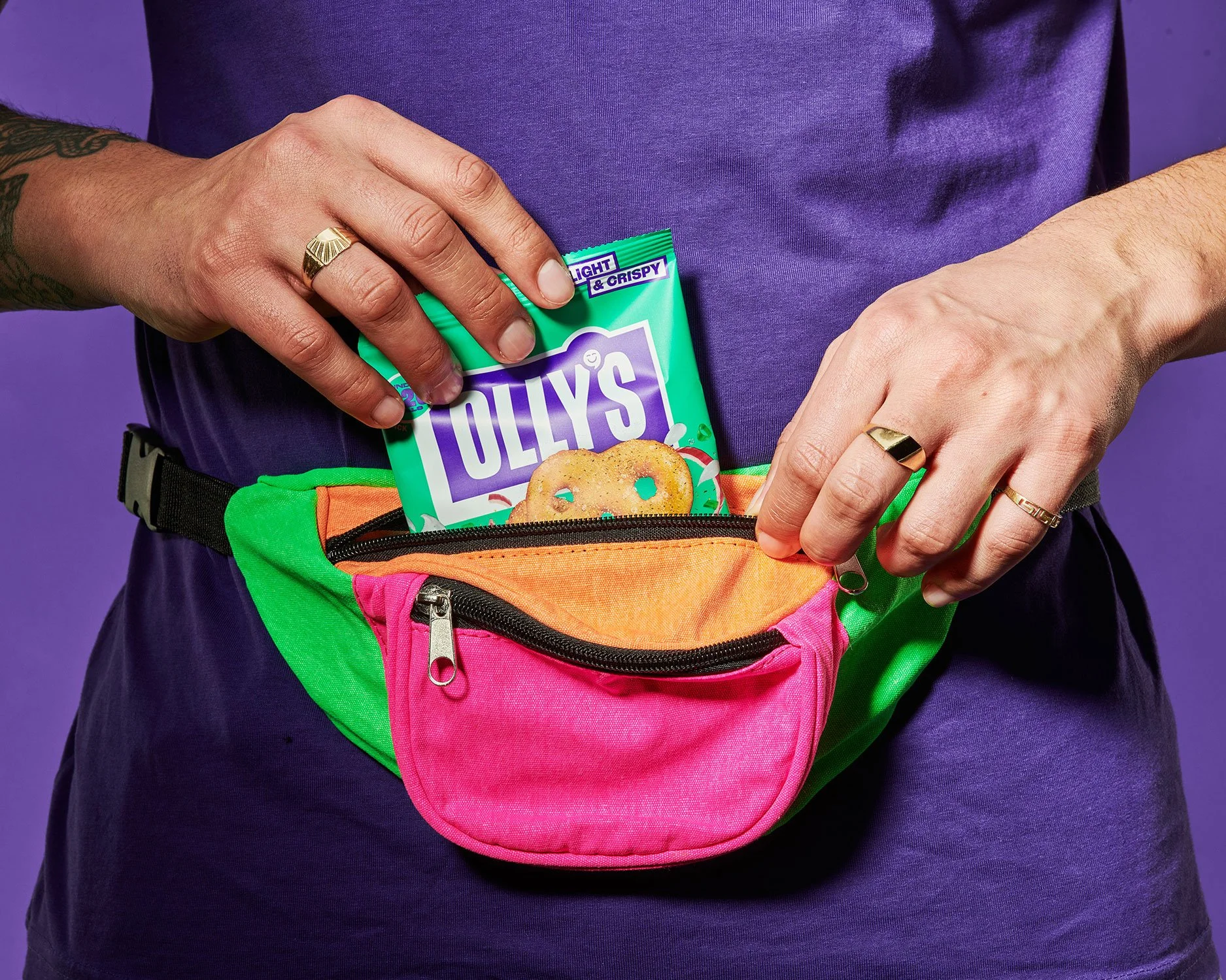 Person wearing a purple shirt with a colorful waist bag, putting a bag of Tollys cookies into the front pocket, against a purple background.