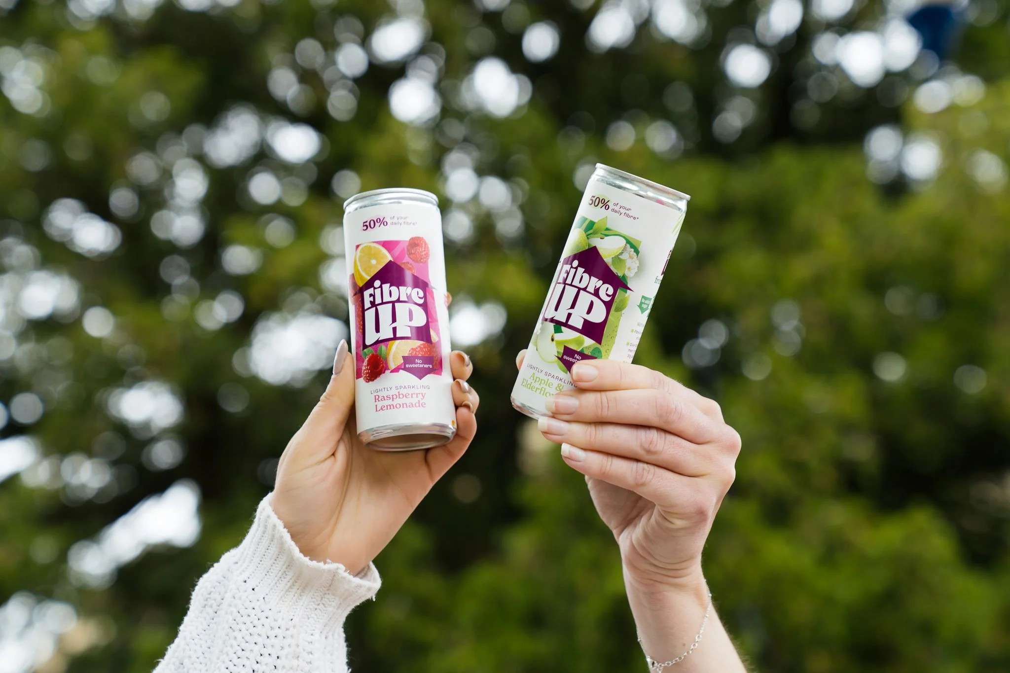 Two hands holding cans of Fibre UP sparkling drinks outdoors with blurred green trees in the background. One can is raspberry lemonade flavor and the other is apple and elderflower flavor.