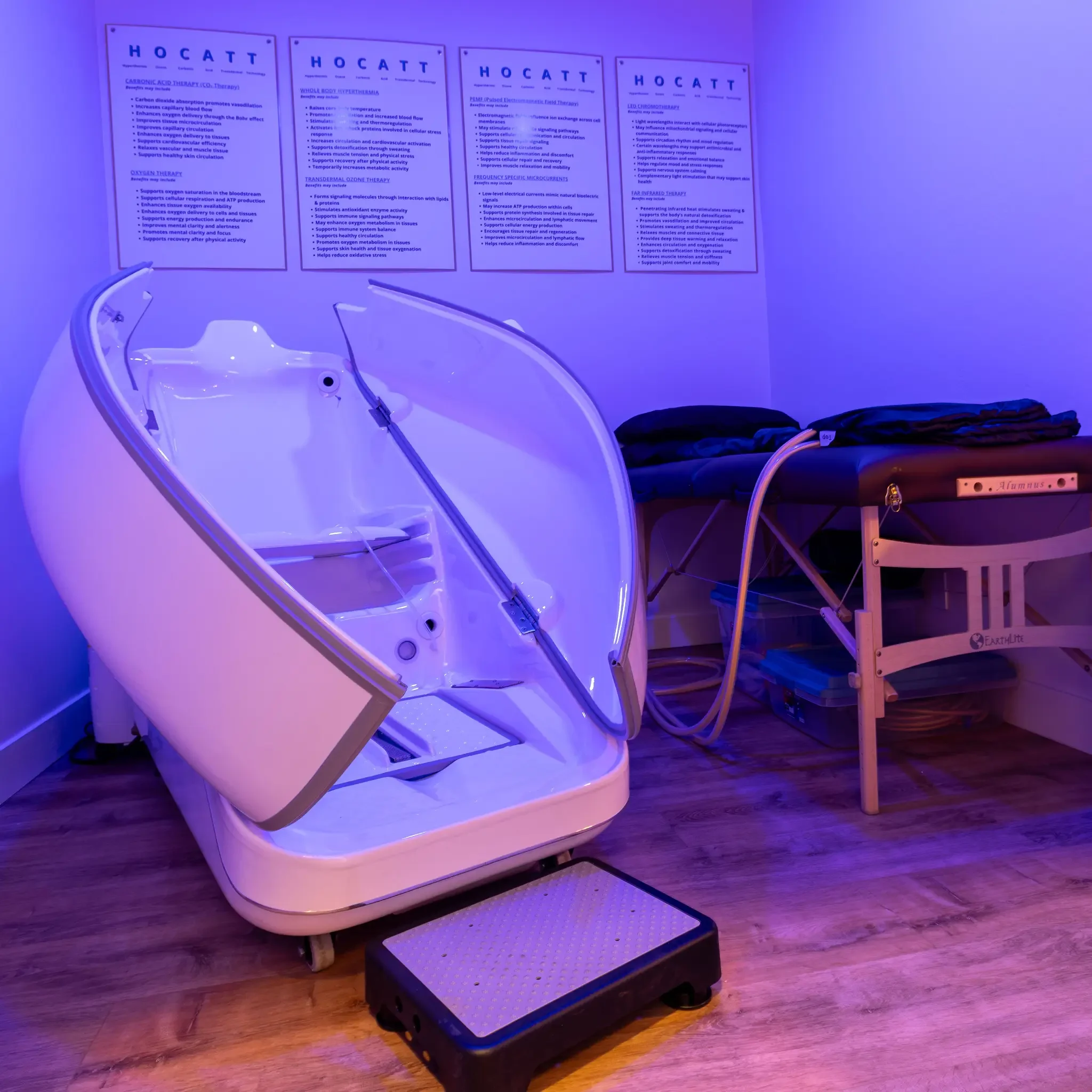 A cryotherapy chamber with a reclining chair inside and posters on the wall detailing various therapy benefits, in a room lit with purple-blue lighting.