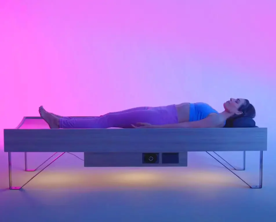 A woman lying on a modern bed with a black pillow beneath her head, in a room with purple and pink lighting.