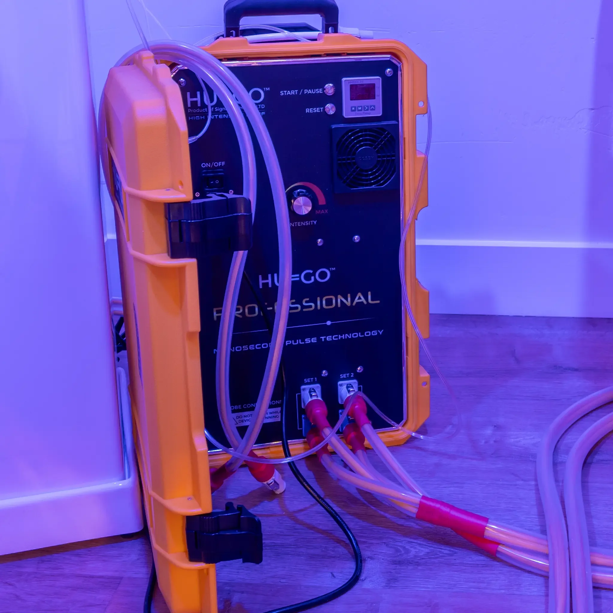 Medical device with tubes and cables attached, labeled 'HUGO.' It is placed on a wooden surface next to a white container, under blue lighting.