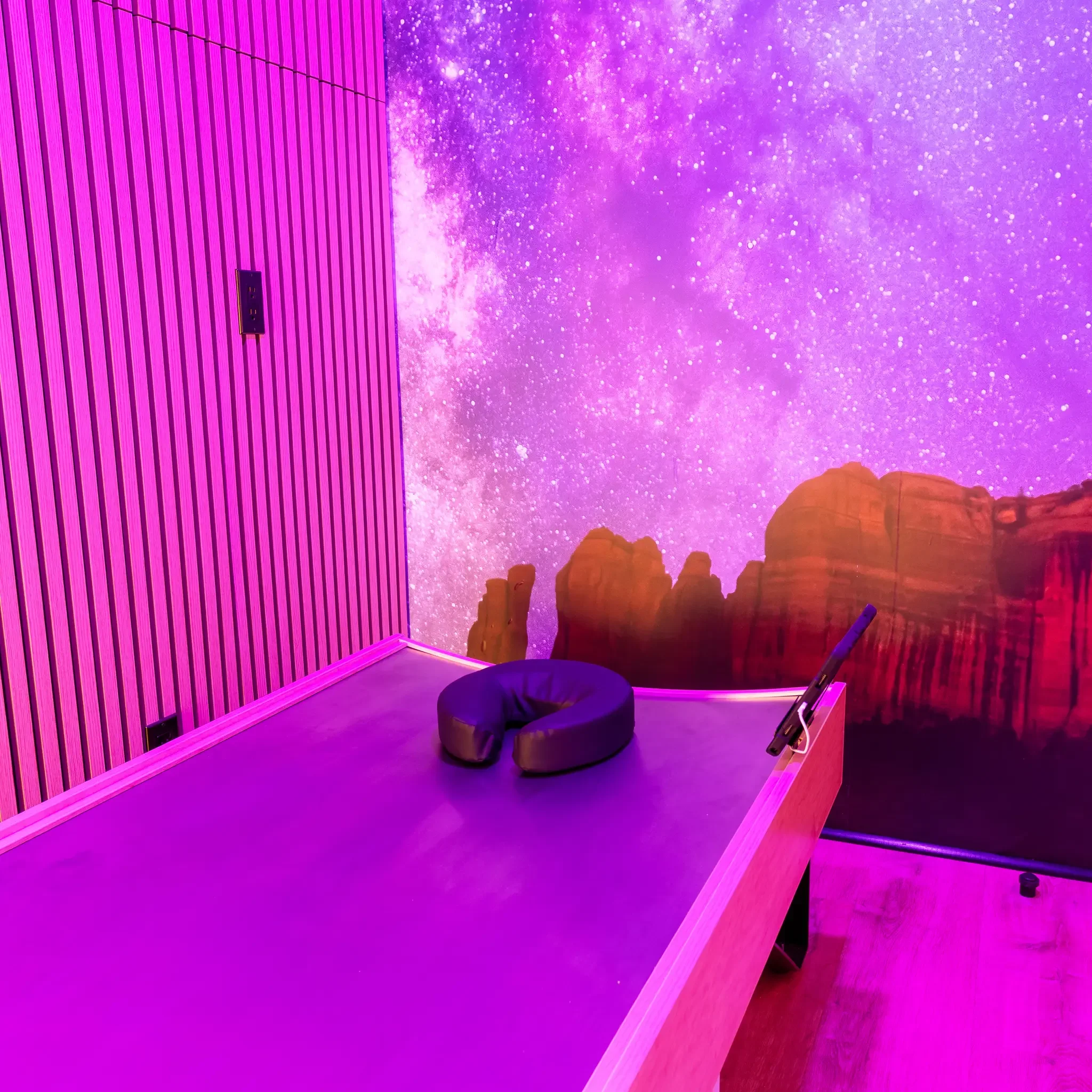 Indoor space with purple and pink lighting, wooden walls, a black bean bag chair, and a wall mural of a starry night sky with red rock formations.