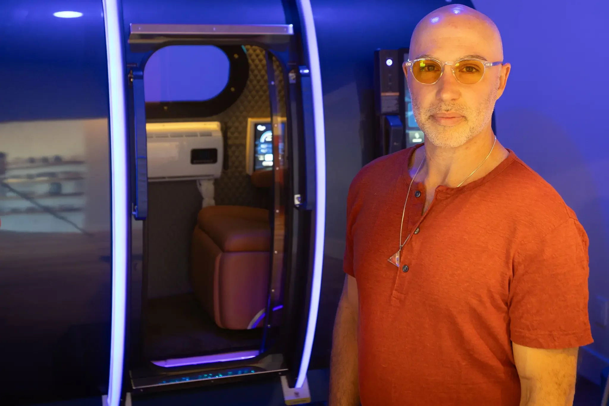 A man with glasses and a reddish-orange shirt standing next to a futuristic-looking capsule or pod with blue and purple neon lighting.