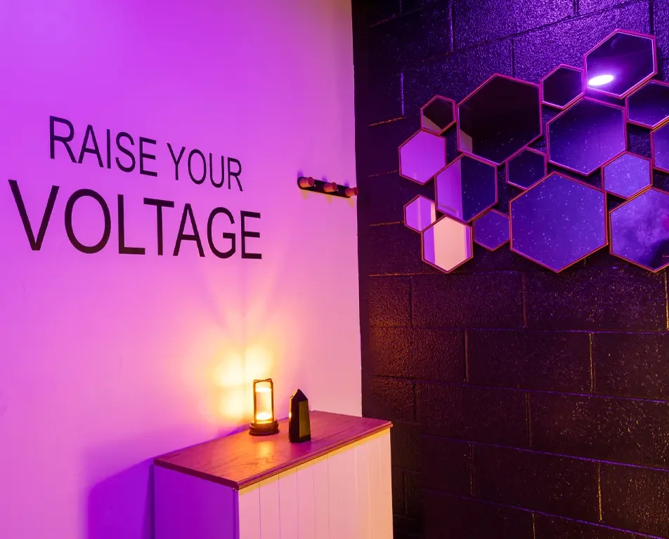 Decorative wall with purple and black hexagon art and the phrase "Raise Your Voltage" on a white wall, with a wooden surface holding a lantern and a small black object, in a dimly lit room with purple and pink lighting.